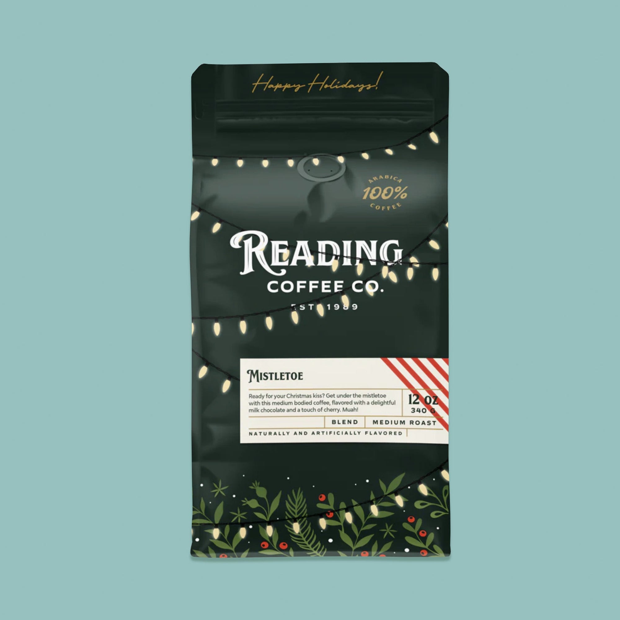 On a sage green background sits a dark green package of coffee. This package is gold, white, and dark green lettering. There are swags of lights and greenery with berries. It says "Happy Holidays!", "Reading Coffee Co. Est. 1989", "Mistletoe Coffee Blend Medium Roast." It is '100% Arabica Coffee. It also says "Ready for your Christmas kiss? Get under the mistletoe wiht this medium bodied coffee, flavored with a delightful milk chocolate and a touch of cherry. Muah!" 12 oz 340g
