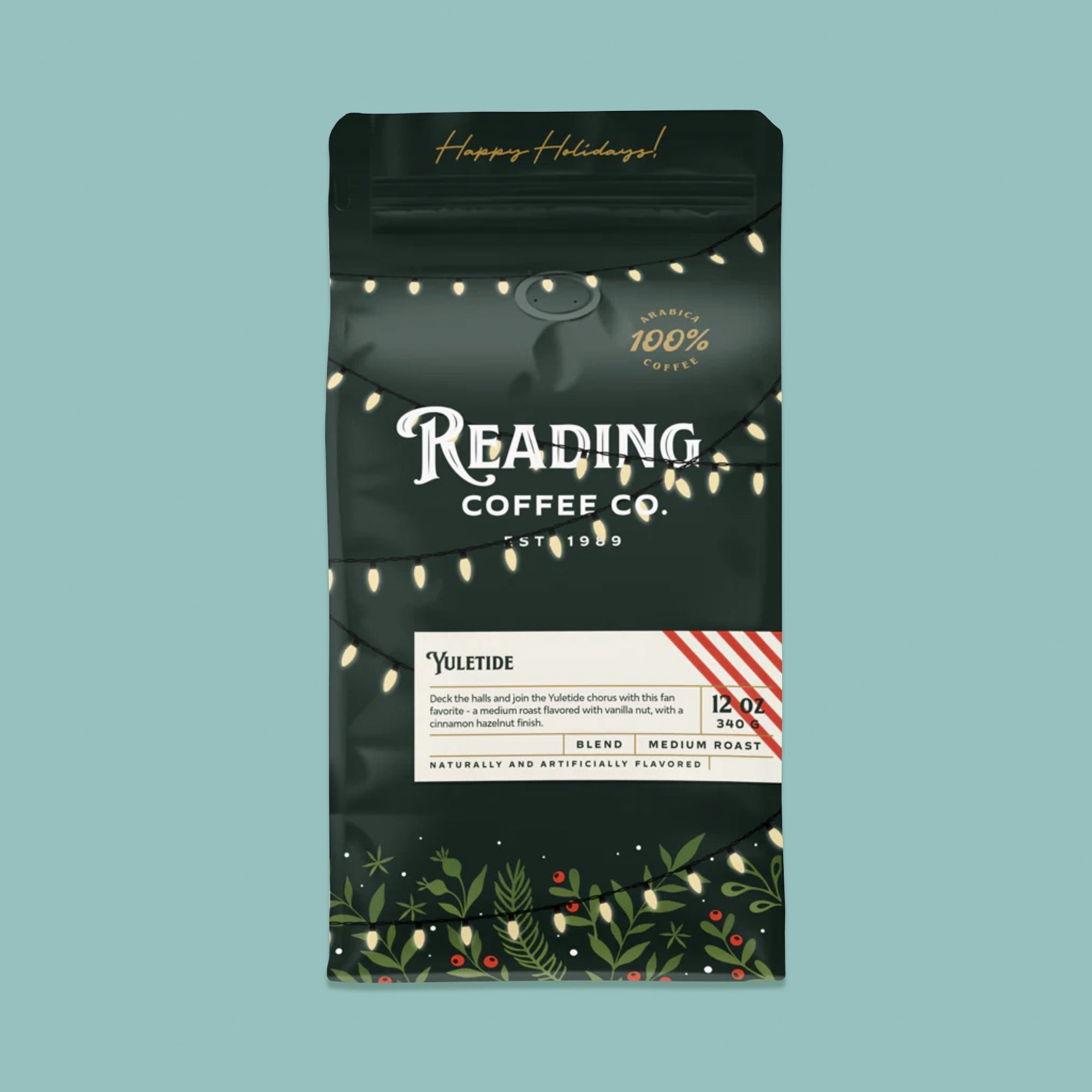 On a sage green background sits a dark green package of coffee. This package is gold, white, and dark green lettering. There are swags of lights and greenery with berries. It says "Happy Holidays!", "Reading Coffee Co. Est. 1989", "Yuletide Blend Medium Roast." It is '100% Arabica Coffee. It also says "Deck the halls and join the Yuletide chorus with this fan favorite - a medium roast flavored with vanilla nut, with a cinnamon hazelnut finish." 12 oz 340g