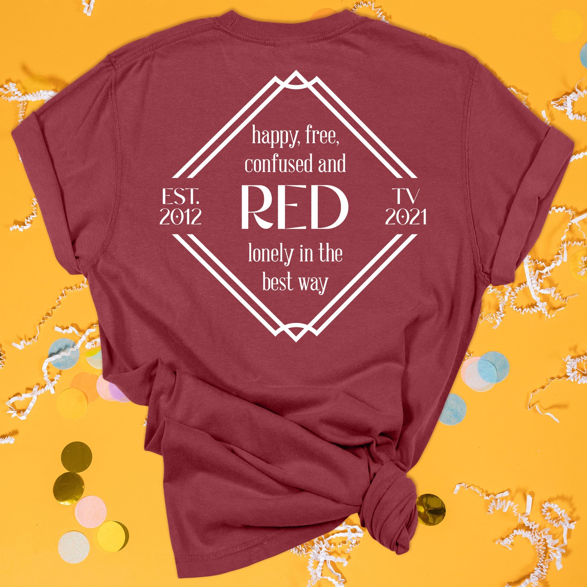 On a sunny mustard background sits the back of a t-shirt with white crinkle and big, colorful confetti scattered around. This Taylor Swift Inspired Reputation tee is dark red with white lettering. It says "happy, free, confused and lonely in the best way" in all lowercase lettering and "RED EST. 2012 TV 2021" in all caps.