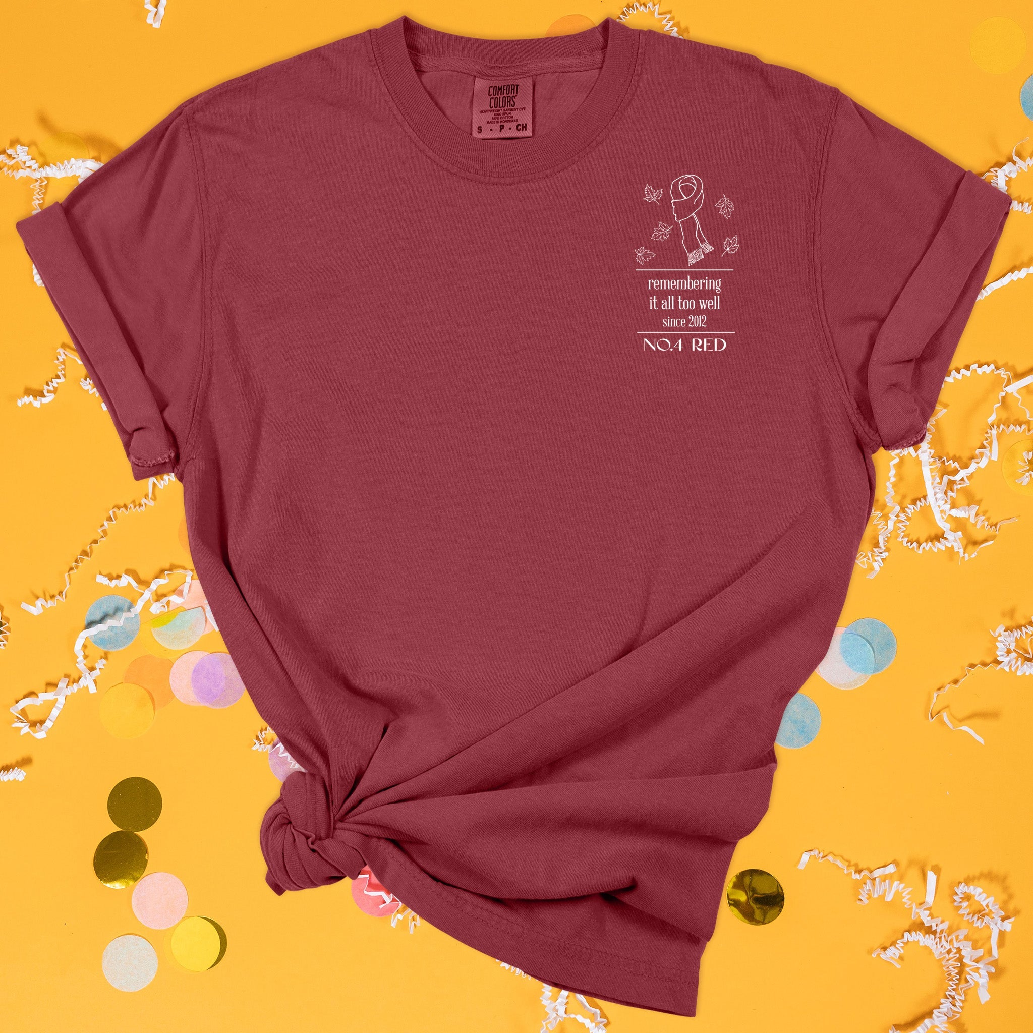 On a sunny mustard background sits the front of a t-shirt with white crinkle and big, colorful confetti scattered around. This Taylor Swift Inspired Reputation tee is dark red with white lettering and illustration. It has a scarf with fall leaves and says "remembering it all too well since 2012" in all lowercase lettering and "NO.4 RED" in all caps.