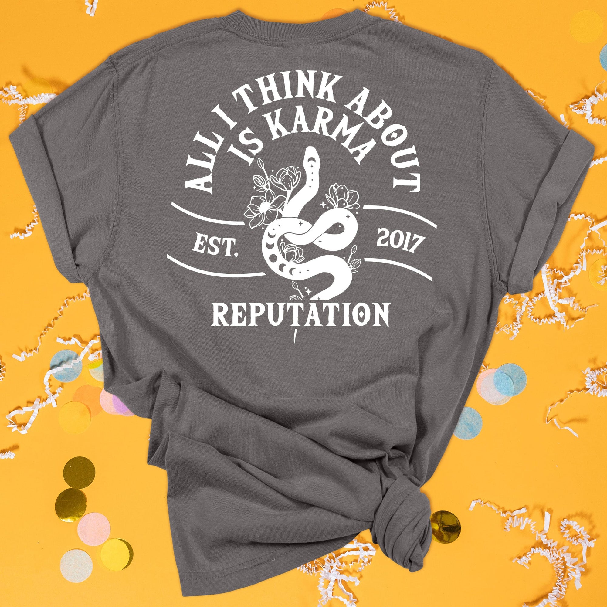 On a sunny mustard background sits the back of a t-shirt with white crinkle and big, colorful confetti scattered around. This Taylor Swift Inspired Reputation tee is dark grey with lettering and illustration. There is an illustration of a snake with flowers and it says "ALL I THINK ABOUT IS KARMA, EST. 2017, REPUTATION" in all caps.
