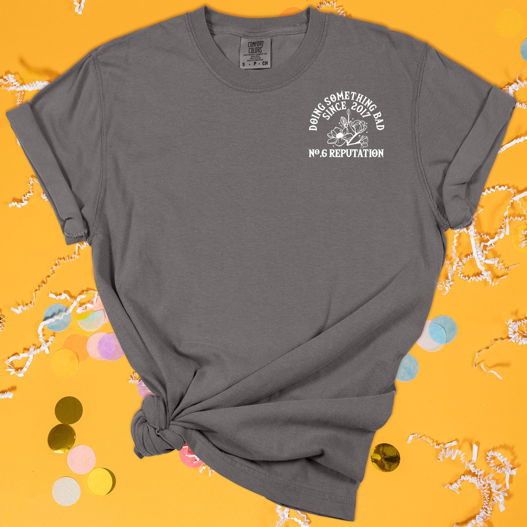 On a sunny mustard background sits the front of a t-shirt with white crinkle and big, colorful confetti scattered around. This Taylor Swift Inspired Reputation tee is dark grey with white lettering and illustration over the top left chest. There are flowers and it says "DOING SOMETHING BAD SINCE 2017, NO.6 REPUTATION" in all caps.