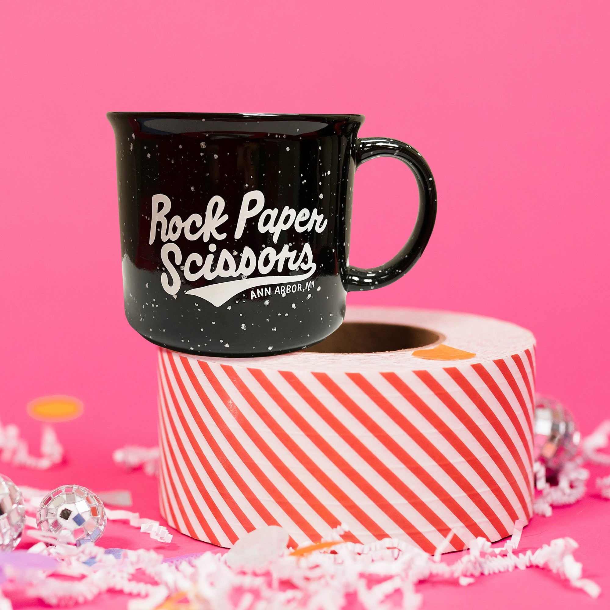 On a hot pink background sits a mug with white crinkle and big, colorful confetti scattered around. There are mini disco balls. This is a black campfire mug with white specks and it says "Rock Paper Scissors" in white, hand lettering.