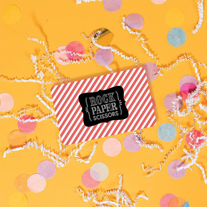 On a sunny mustard background is a gift card with white crinkle and big, colorful confetti scattered around. This E-gift card has red and white stripes with a black and white Rock Paper Scissors logo on top. 