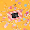 On a sunny mustard background is a gift card with with white crinkle and big, colorful confetti scattered around. This gift card has red and white stripes with a black and white Rock Paper Scissors logo on top. 