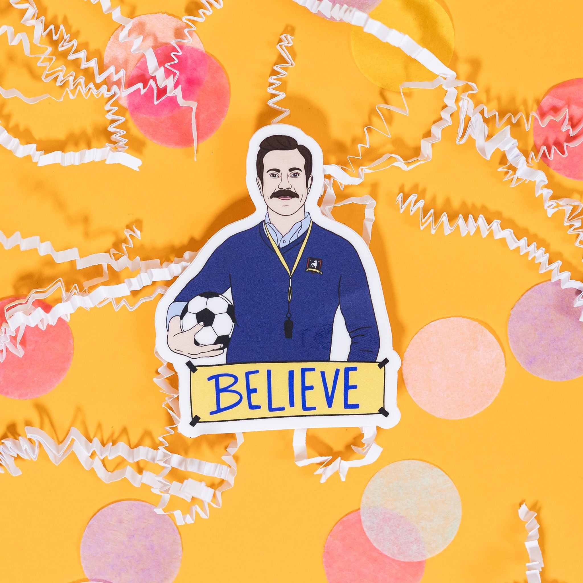 On a sunny mustard background sits a sticker with white crinkle and big, colorful confetti scattered around. This sticker is an illustration of Ted Lasso holding a futbol with a sign at the bottom that says "BELIEVE" in blue handwritten lettering. The colors are in navy, yellow, blue, and light blue with black and white. 2"-3"