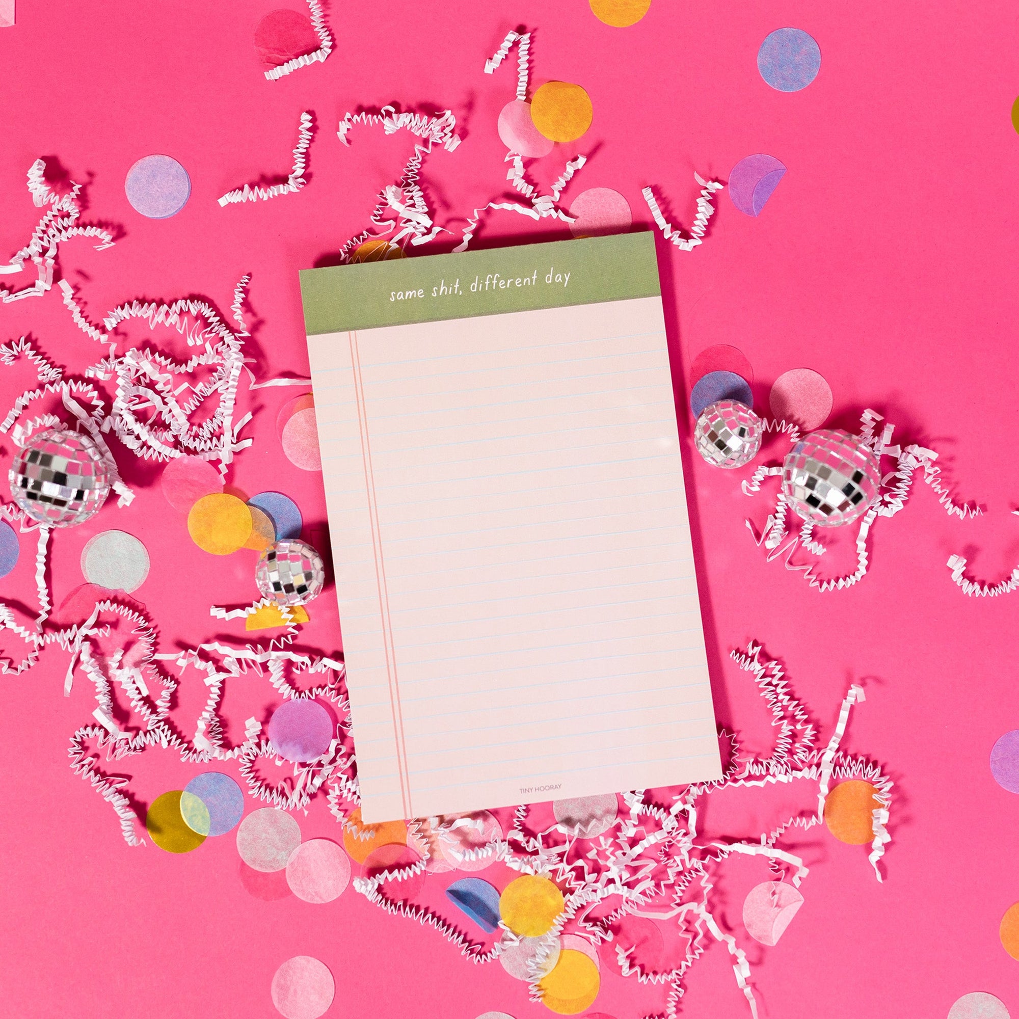 On a hot pink background sits a light pink notepad with white crinkle and big, colorful confetti scattered around. There are mini disco balls. It says "same shit, different day" at the top and has blue lines.