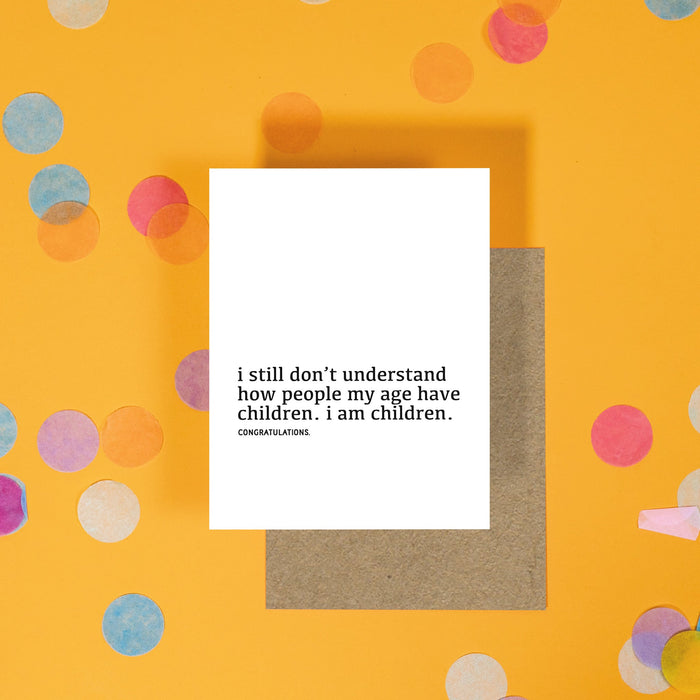 On a sunny mustard background is a greeting card and envelope with white crinkle and big, colorful confetti scattered around. The white greeting card says "I still don't understand how people my age have children. I am children. congratulations." in a black, lower case serif font. A kraft envelope sits under the card.
