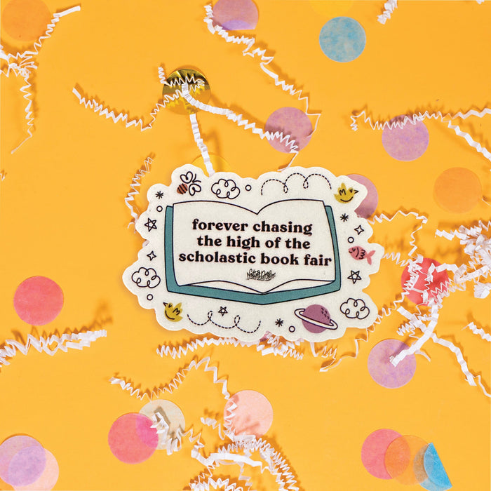 On a sunny mustard background sits a sticker with white crinkle and big, colorful confetti scattered around. This colorful sticker has an illustration of an open book and handdrawn doodles. It says "forever chasing the high of the scholastic book fair" in black, thick serif lettering.