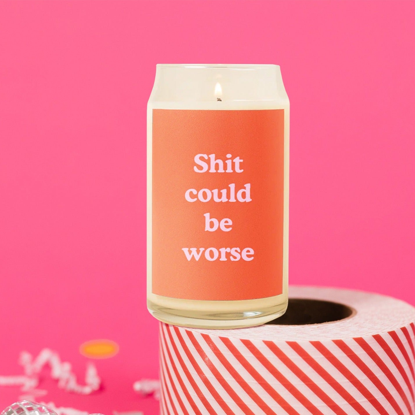 On a hot pink background sits a candle with white crinkle and big, colorful confetti scattered around. The candle is lit and has an orange label on the front that says "Shit could be worse" in a bubblegum pink, thick serif font.