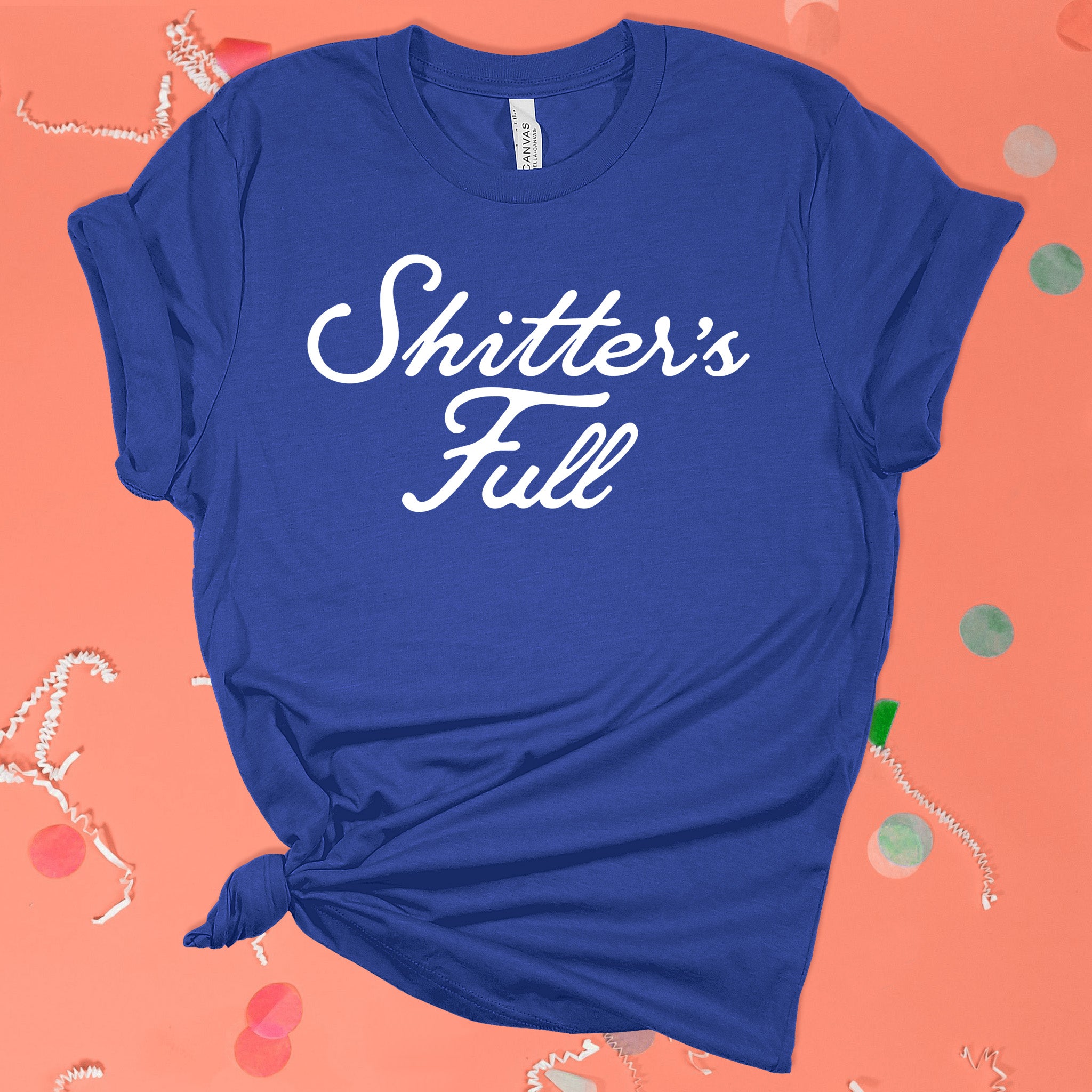 On a pink coral background sits the front of a t-shirt with white crinkle and big, colorful confetti scattered around. This Christmas Vacation Inspired crewneck cobalt blue t-shirt has white script lettering and it says "Shitter's Full."