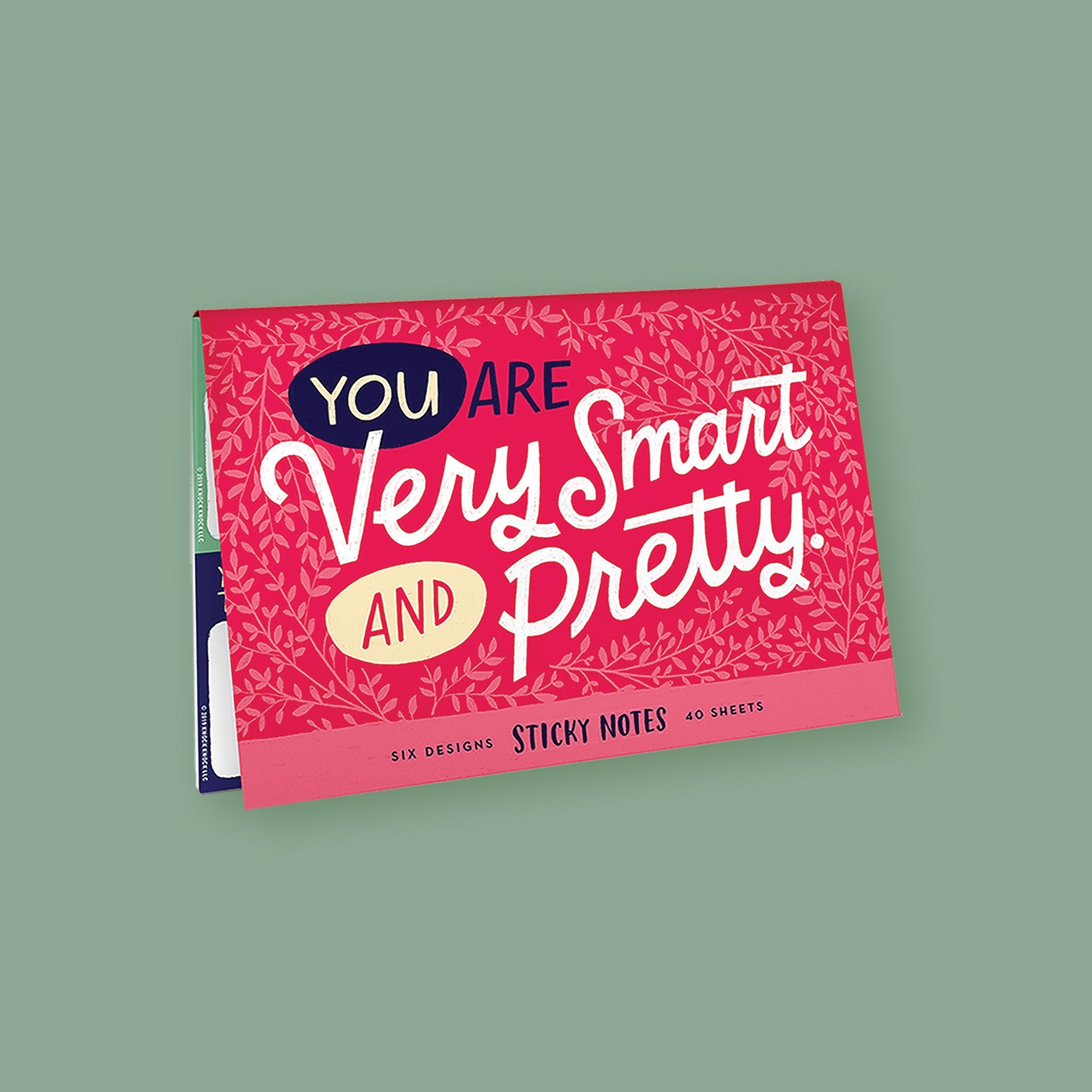 On a moss green background sits a packet. This is a close-up of a cherry pink packet of sticky notes that has light pink illustrations of branches with leaves. It says "YOU ARE Very Smart AND Pretty" in handwritten lettering. The colors of the lettering are navy, light yellow, and white. On the bottom it says "SIX DESIGNS STICKY NOTES 40 SHEETS" in navy, handwritten lettering.