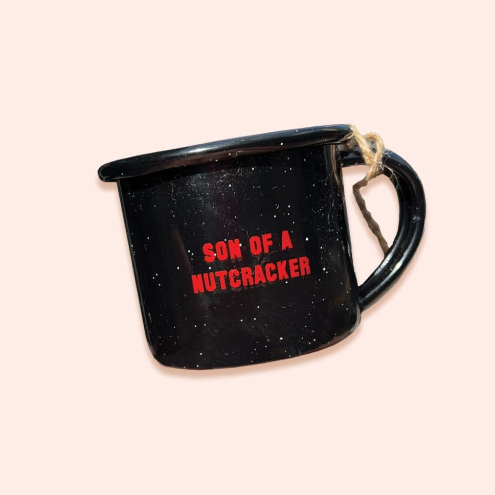 On a light pink background sits a black mini campfire mug with white specks. It says "SON OF A NUTCRACKER" in red collegiate lettering. It has twine on the handle.