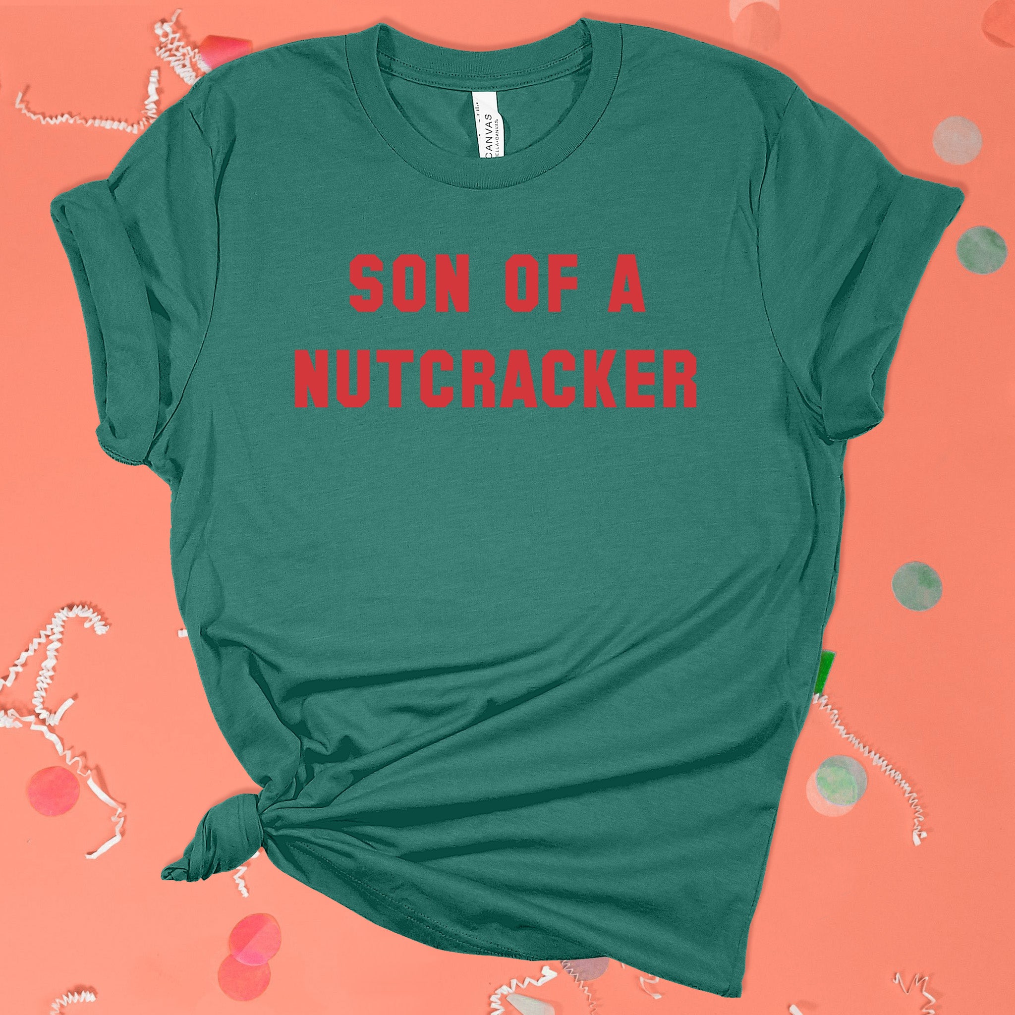 On a pink coral background sits the front of a t-shirt with white crinkle and big, colorful confetti scattered around. This Elf Inspired crewneck t-shirt is dark green with red collegiate lettering. It says "SON OF A NUTCRACKER."