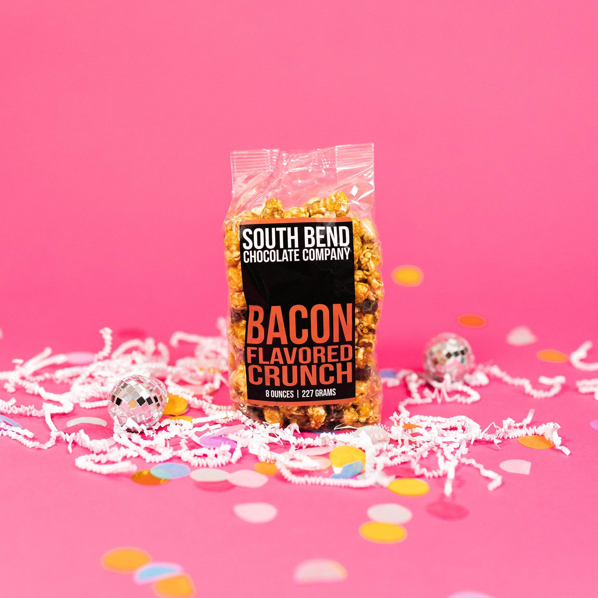On a bubblegum pink background sits a bag with white crinkle and big, colorful confetti scattered around. There are mini disco balls. This package has a black label and it says in white all caps lettering "SOUTH BEND CHOCOLATE COMPANY" and in orange all caps lettering "BACON FLAVORED CRUNCH." 8 ounces | 227 grams