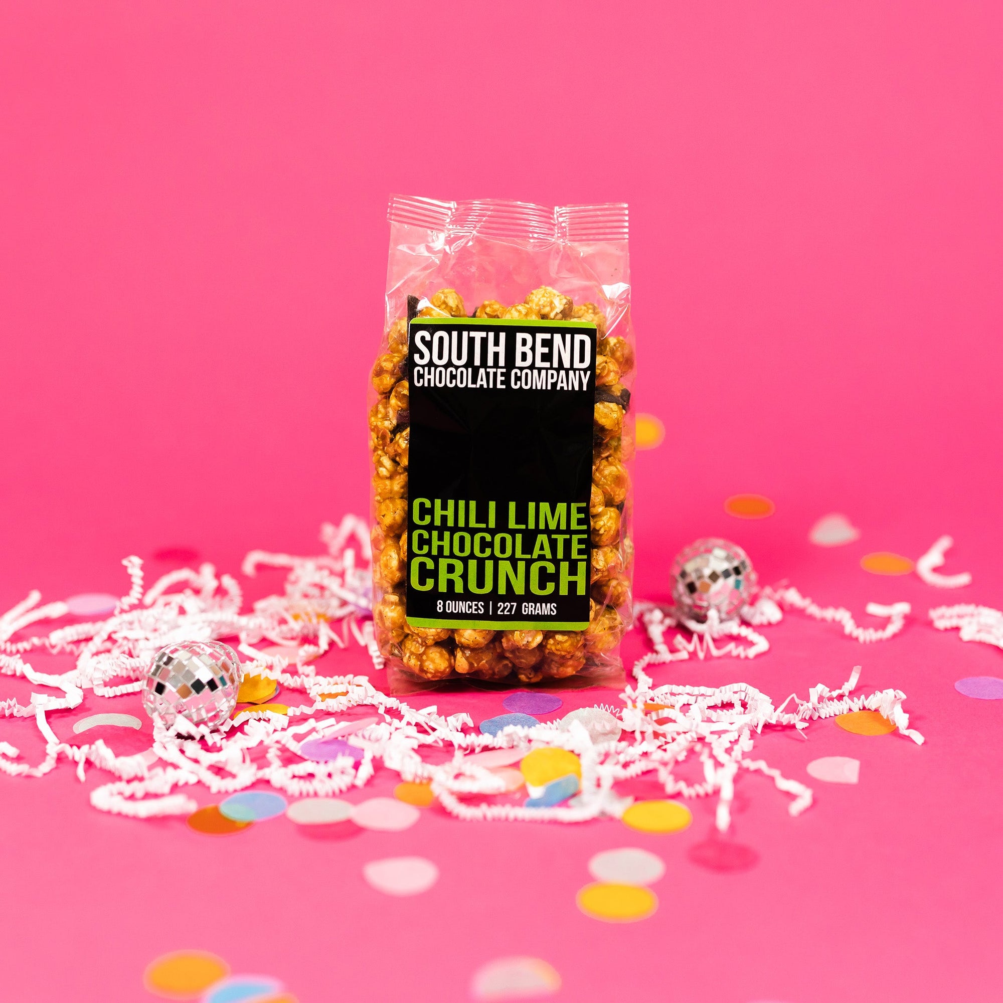 On a bubblegum pink background sits a bag with white crinkle and big, colorful confetti scattered around. There are mini disco balls. This package has a black label and it says in white all caps lettering "SOUTH BEND CHOCOLATE COMPANY" and in lime green all caps lettering "CHILI LIME CHOCOLATE CRUNCH." 8 ounces | 227 grams