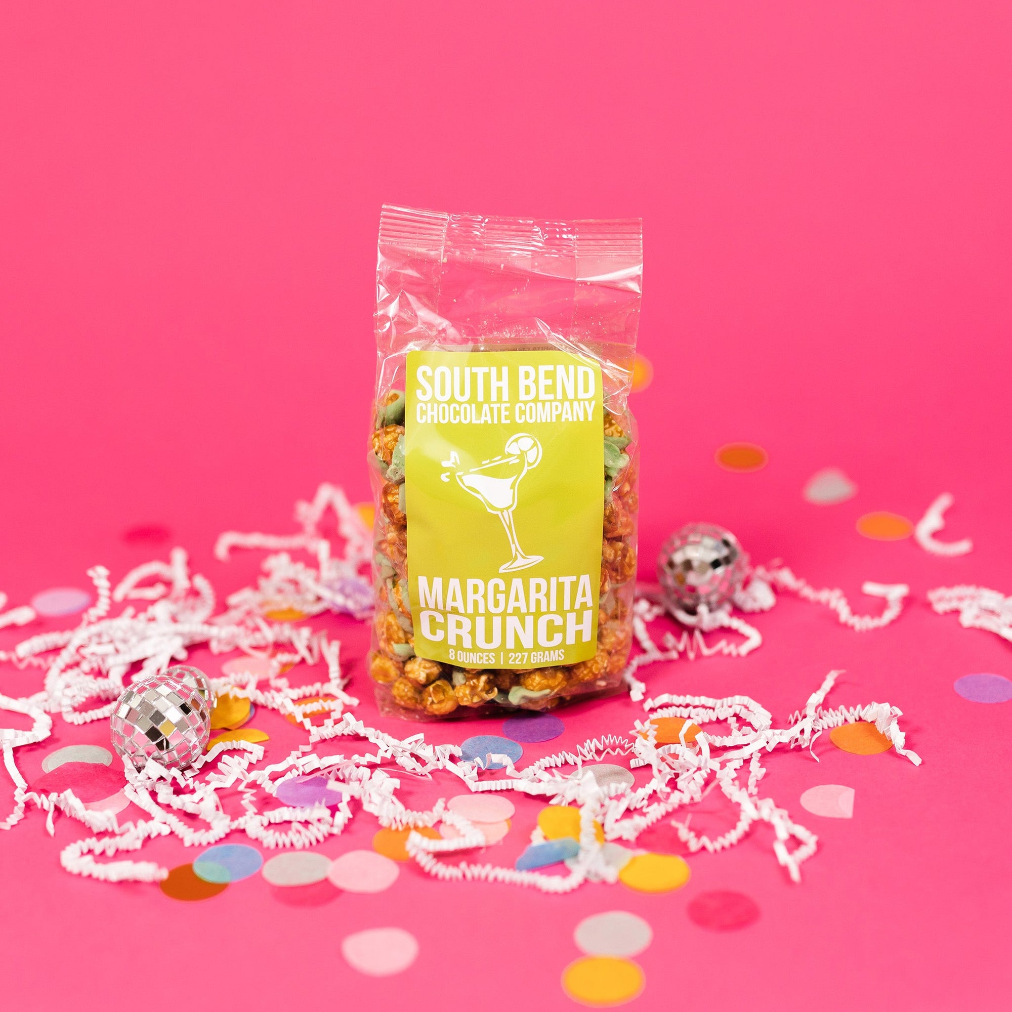 On a bubblegum pink background sits a bag with white crinkle and big, colorful confetti scattered around. There are mini disco balls. This package has a lime green label and it says in white all caps lettering "SOUTH BEND CHOCOLATE COMPANY" and "MARGARITA CRUNCH." Ther is an illustration of a white margarita. 8 ounces | 227 grams