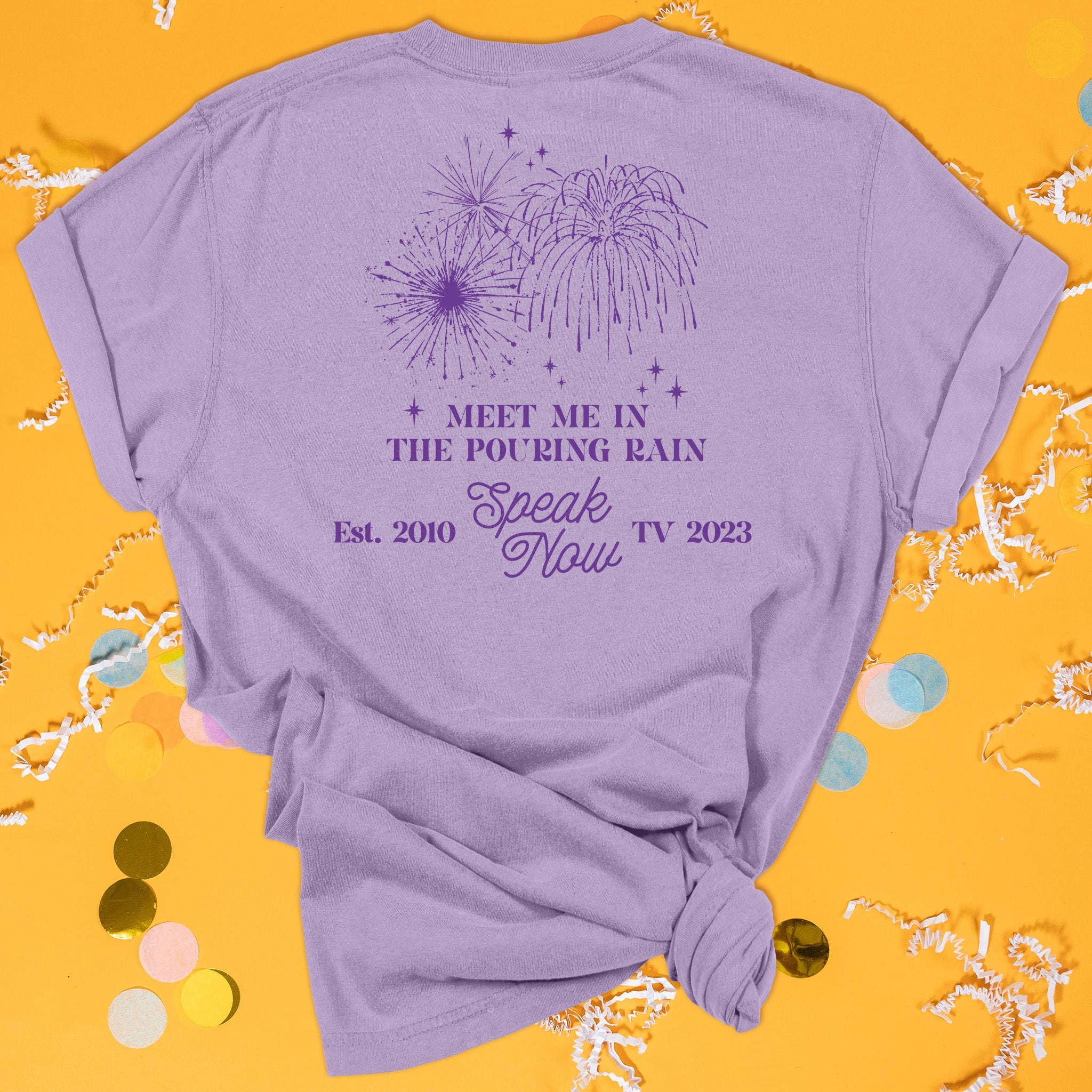 On a sunny mustard background sits the back of a t-shirt with white crinkle and big, colorful confetti scattered around. This Taylor Swift Inspired Reputation tee is dark lavender with dark purple lettering and an illustration of fireworks and sparkle stars. It says "MEET ME IN THE POURING RAIN" in all caps and "Speak Now" in script lettering and "Est. 2010 TV 2023."