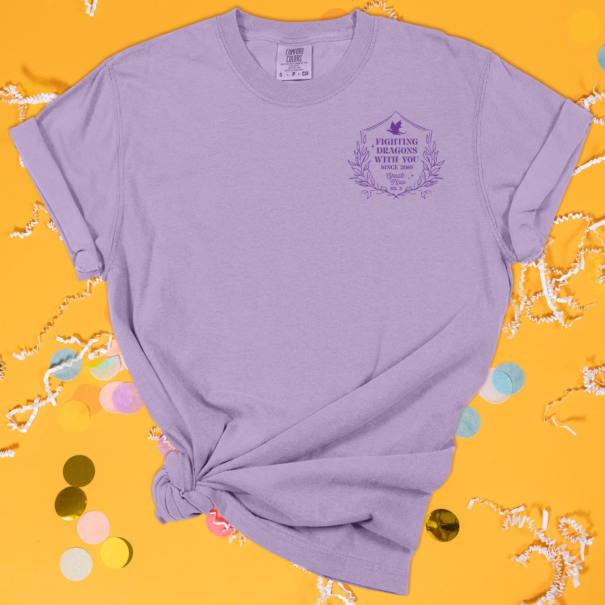 On a sunny mustard background sits the front of a t-shirt with white crinkle and big, colorful confetti scattered around. This Taylor Swift Inspired Reputation tee is dark lavender with dark purple lettering and an illustration of a crest with leaves and a dragon over the left chest area. It says "FIGHTING DRAGONS WITH YOU SINCE 2010" in all caps and "Speak Now" in script lettering and "NO.3."