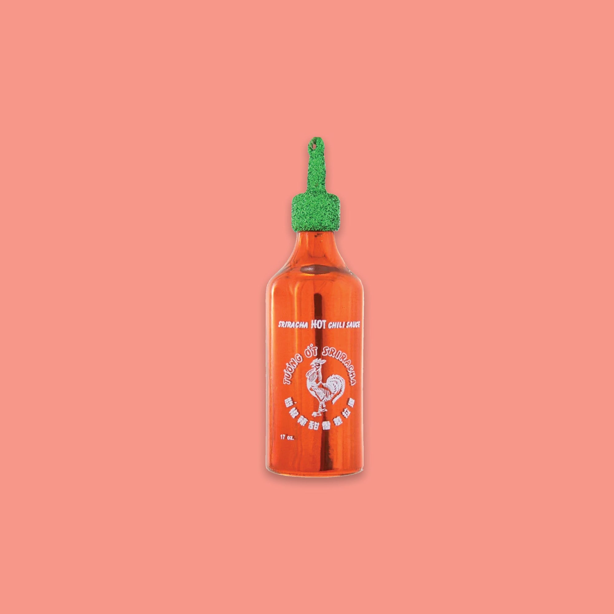 On a coral pink background sits a sriracha inspired ornament. This orange glass ornament has a green glitter top and it says in white on the bottle "SRIRACHA HOT CHILI SAUCE." It has chinese writing and a rooster.