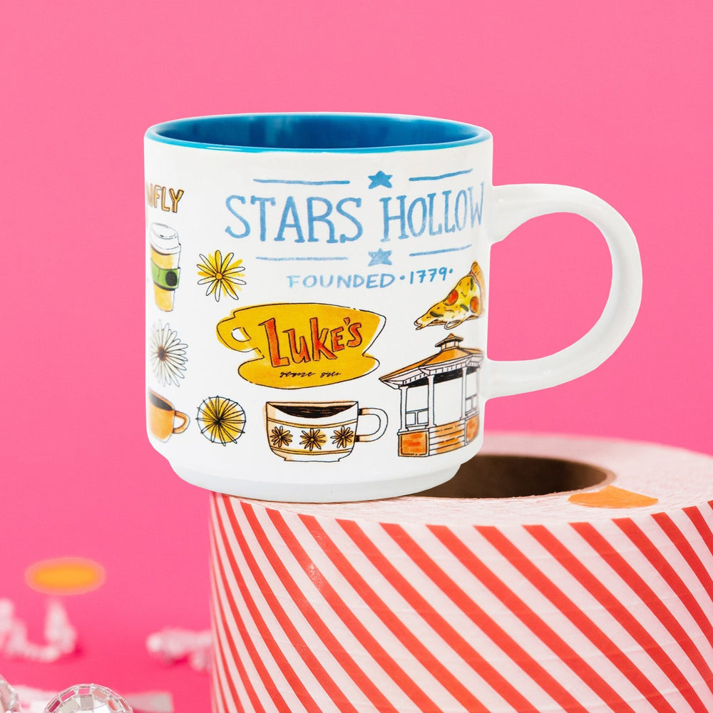 On a hot pink background sits a candle on a red and white packing tape with colorful confetti and white crinkle scattered around. This Gilmore Girls inspired mug has illustrations of Luke's Pizza, the gazebo , sunflowers and coffee cups. There are illustrations of Dragon Fly Inn for this 11 ounce white mug. Illustrations are in bright colors.