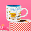 On a hot pink background sits a candle on a red and white packing tape with colorful confetti and white crinkle scattered around. This Gilmore Girls inspired mug has illustrations of Luke's Pizza, the gazebo , sunflowers and coffee cups. There are illustrations of Dragon Fly Inn for this 11 ounce white mug. Illustrations are in bright colors.