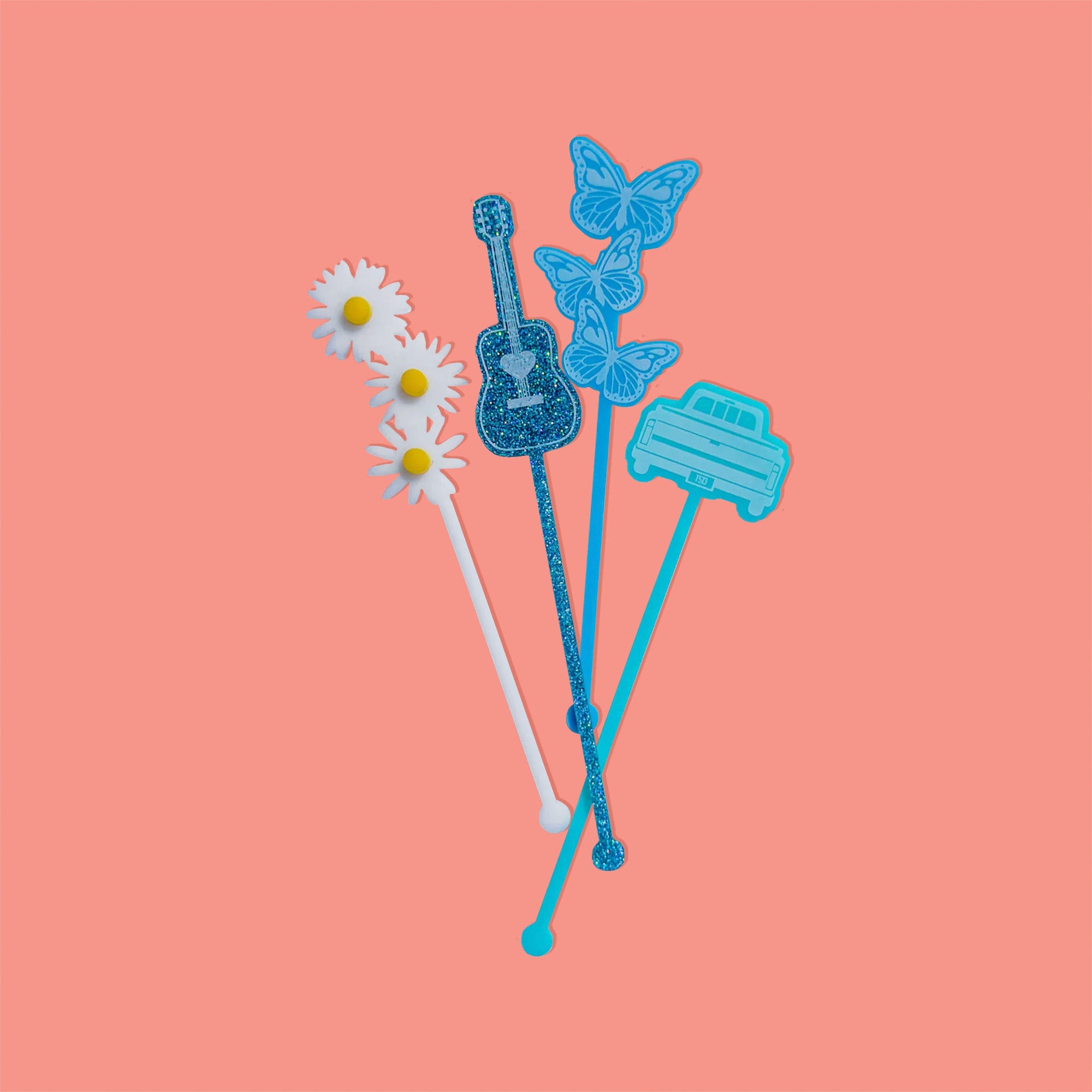 On a coral pink background sits acrylic stirrers. This Taylor Swift inspired set of 4 stirrers includes a white with yellow opaque daisies, a teal glitter acoustic guitar, a trio of laguna opague butterflies, and a turquoise opaque pickup truck.