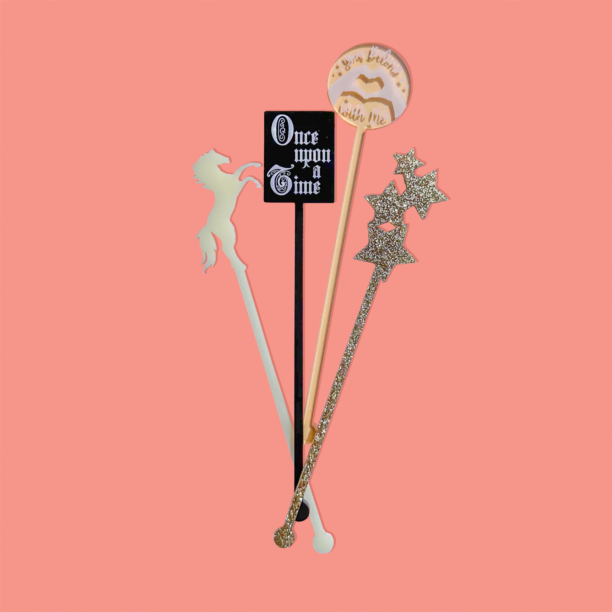 On a coral pink background sits acrylic stirrers. This Taylor Swift inspired set of 4 stirrers includes an ivory horse, gold transparent heart hands, gold glitter stars, and a black opaque "Once upon a Time" fairytale book.