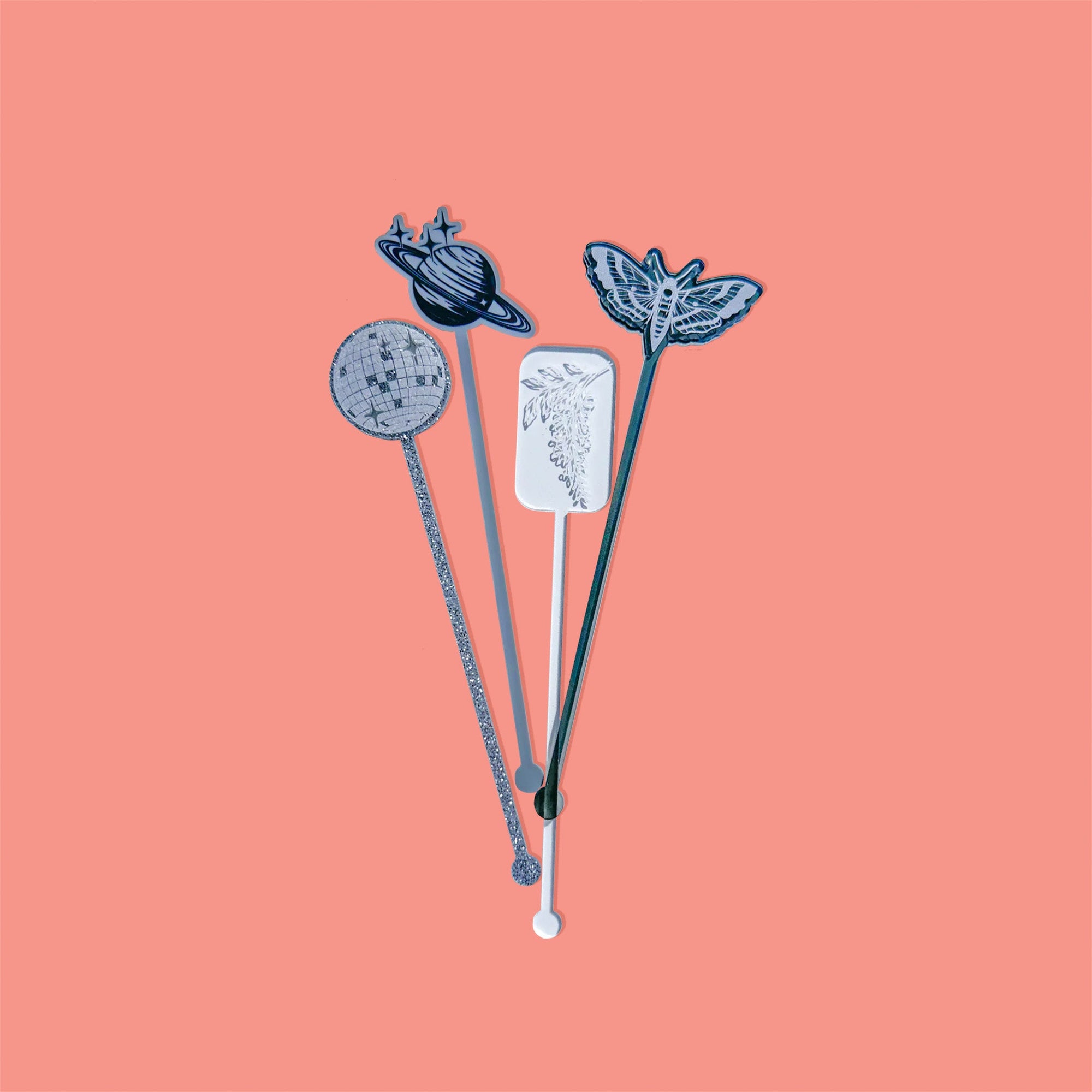 On a coral pink background sits acrylic stirrers. This Taylor Swift inspired set of 4 stirrers includes a silver glitter mirrorball, a clear transparent rectangle with white wisteria, a gray opaque Saturn sketch, and a gray smoke moth.