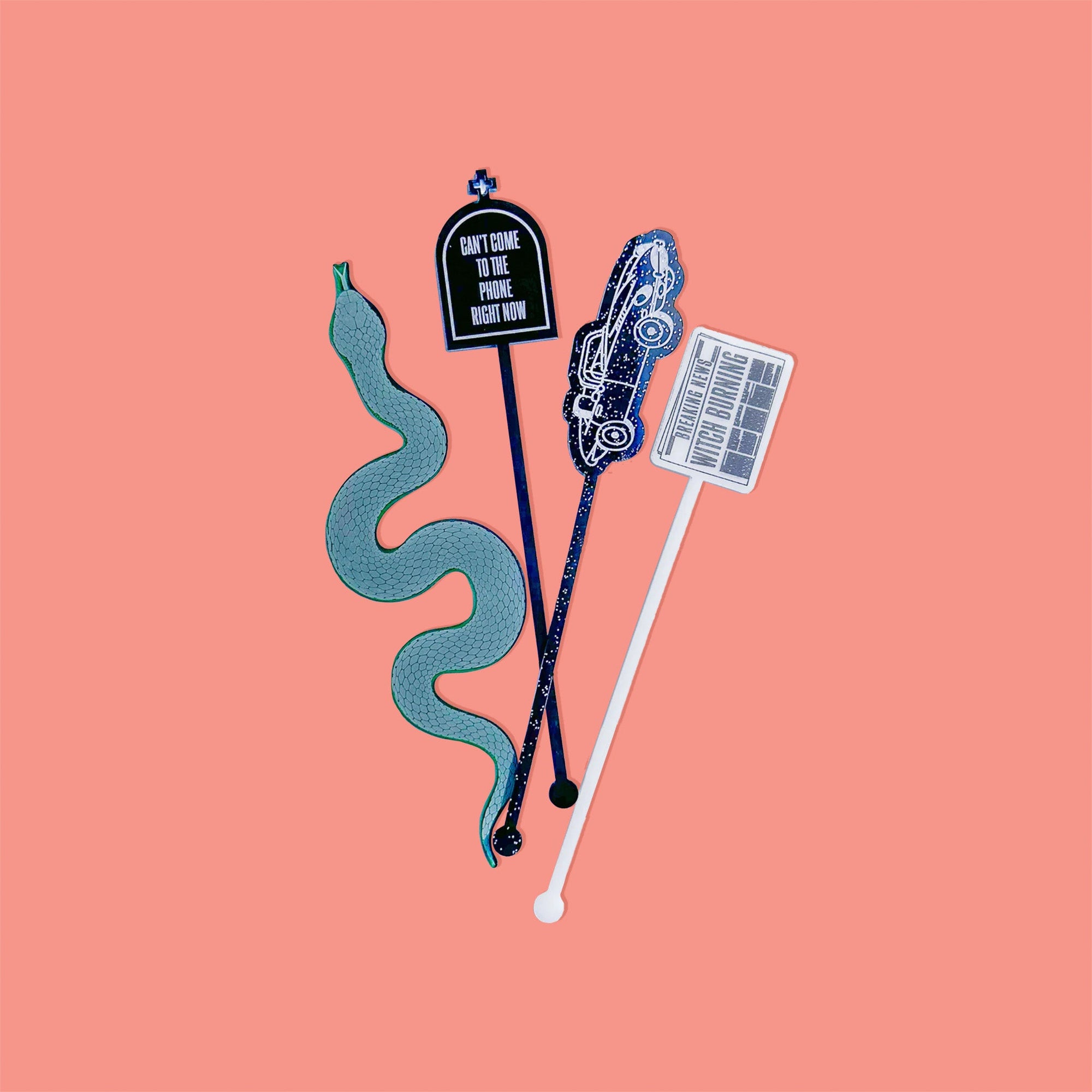 On a coral pink background sits acrylic stirrers. This Taylor Swift inspired set of 4 stirrers includes a dark green transparent snake, a black opaque tombstone, a white opague newspaper, and a starlight glitter getaway car.