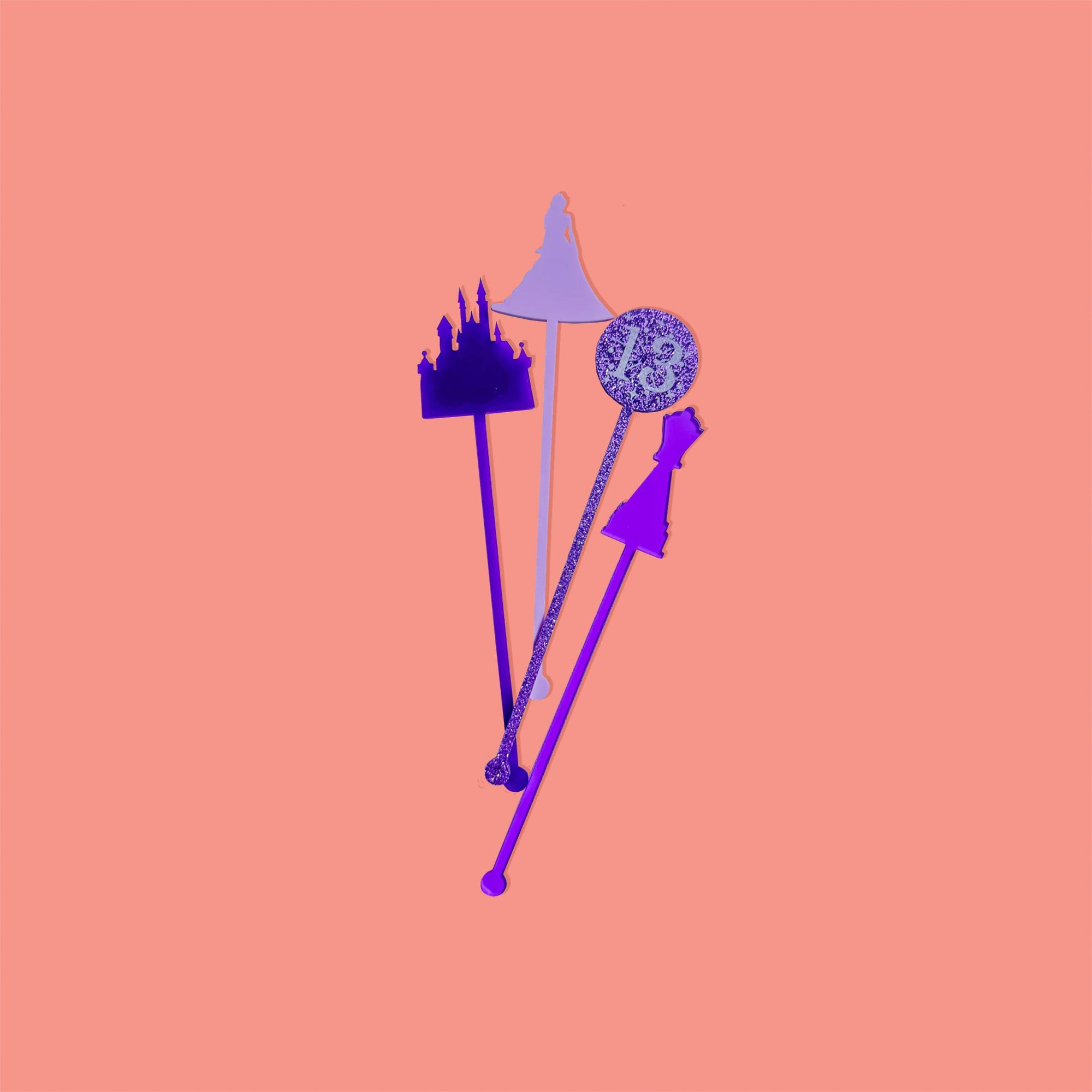 On a coral pink background sits acrylic stirrers. This Taylor Swift inspired set of 4 stirrers includes a purple opaque castle, a purple transparent chess piece, a purple glitter 13 sparkler, and a lilac opaque enchanted dress.