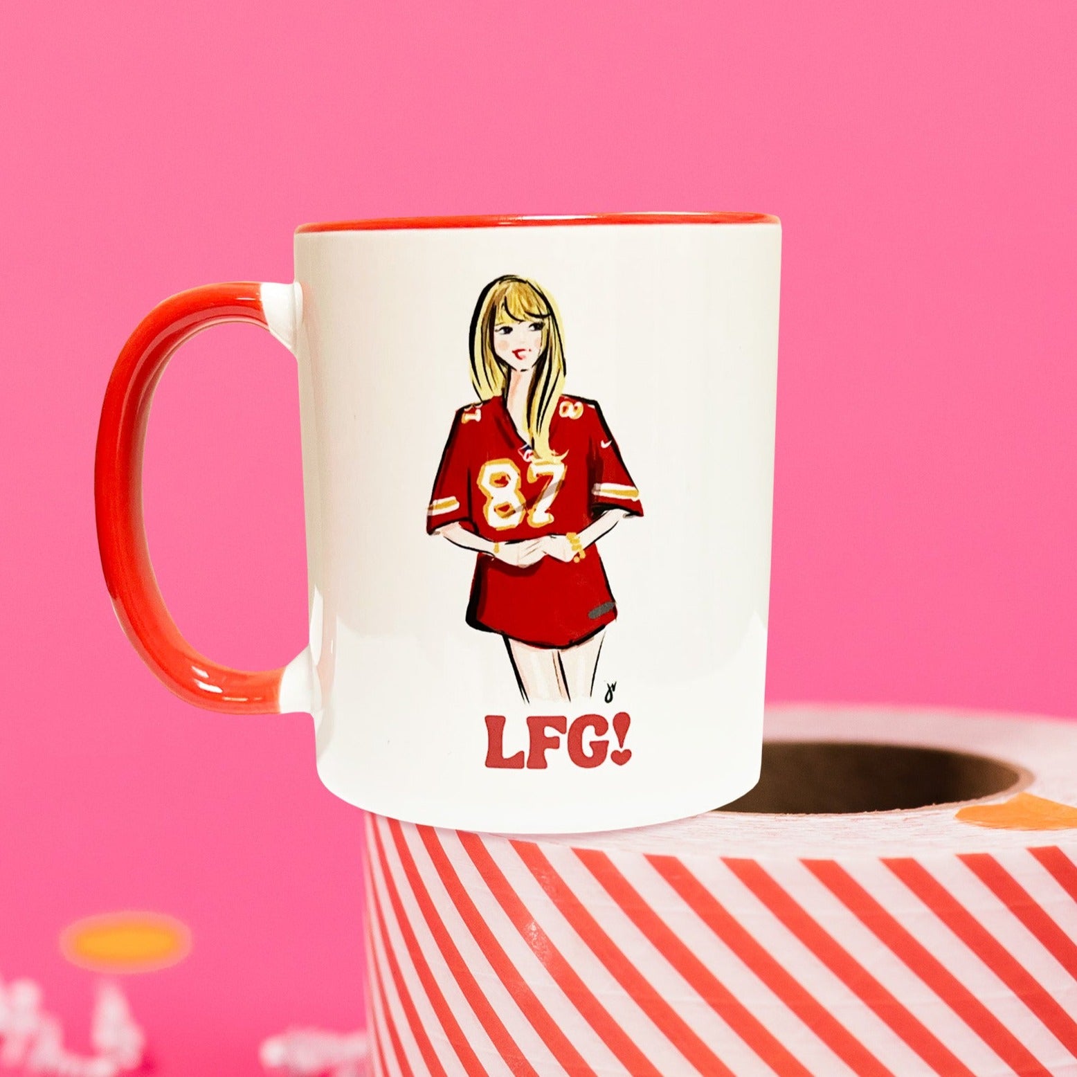 On a hot pink background sits a mug on top of a red and white striped packing tape with white crinkle and big, colorful confetti scattered around. There are mini disco balls. This Taylor Swift inspired mug has an illustration of Taylor Swift wearing a Kansa City Chiefs jersey with the number 87 on it. It has a red handle and rim.
