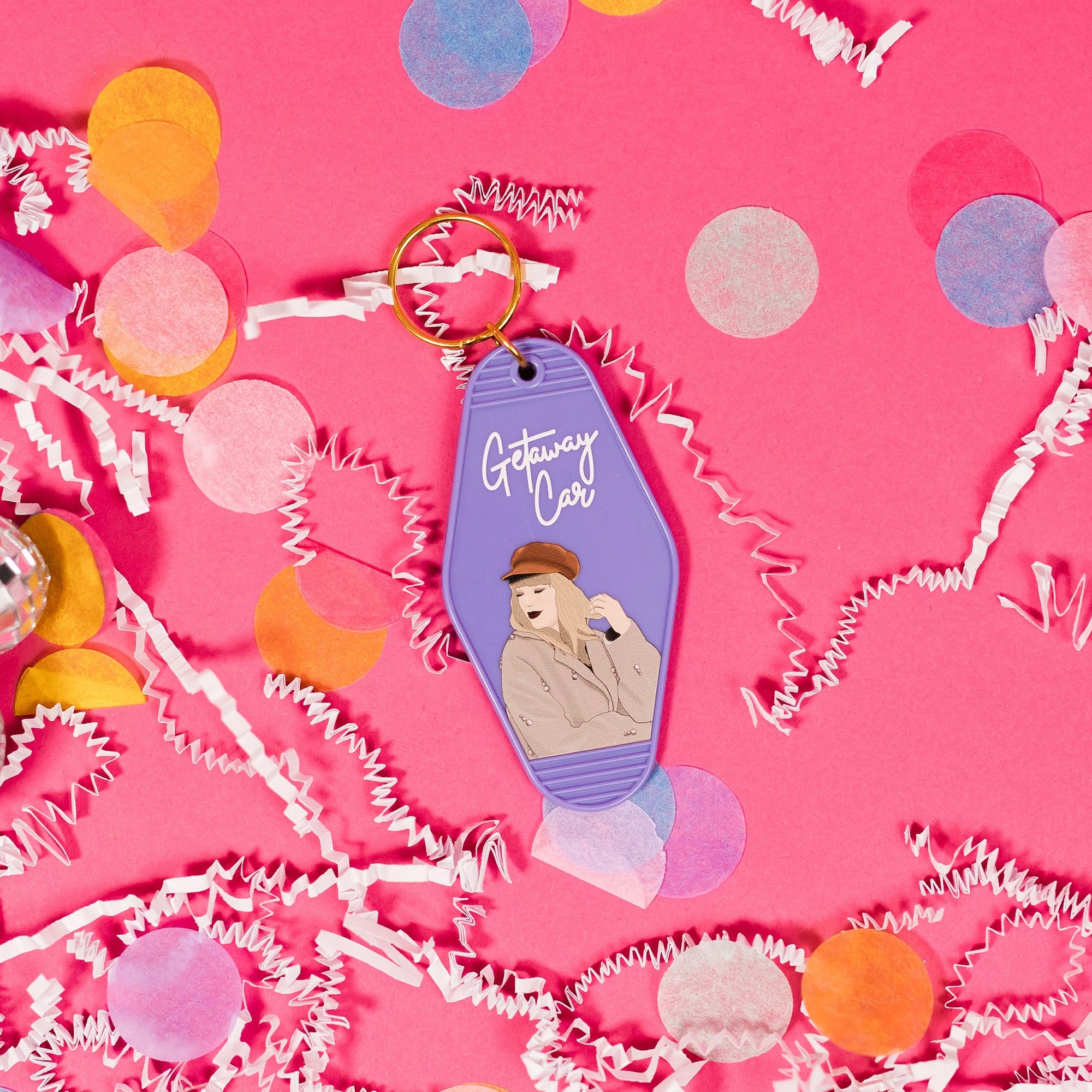 On a hot pink background sits a keychain with white crinkle and big, colorful confetti scattered around. There are mini disco balls. This Taylor Swift inspired vintage, motel keychain is lavender with an illustration of Taylor Swift wearing a hat and coat. In a white, handwritten lettering script it says "Getaway Car".