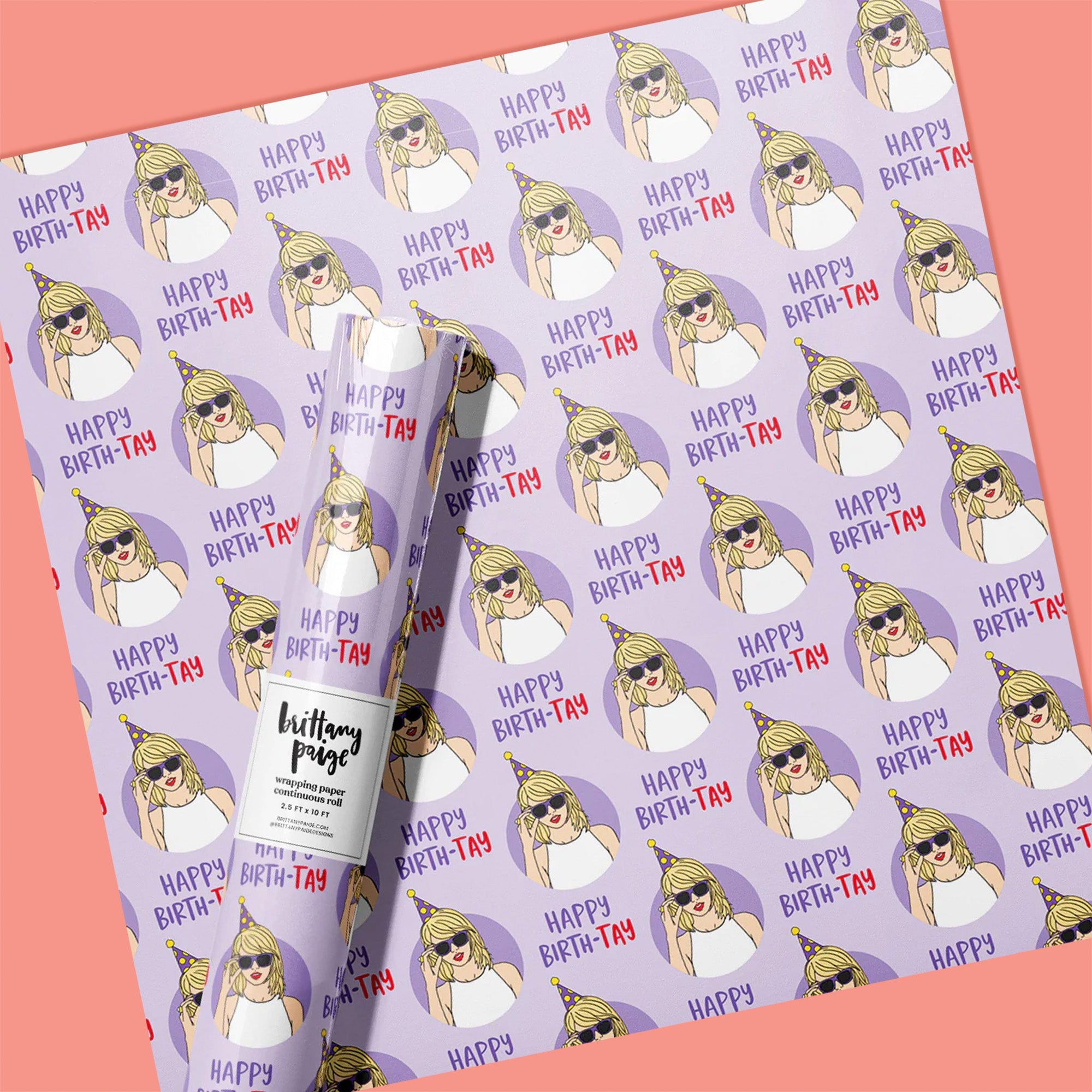 On a coral pink background sits a gift wrap sheet and a roll. This Taylor Swift inspired gift wrap is light purple with darker purple circles with illustrations of Taylor Swift wearing a party hat and sunglasses. It says "Happy Birth-Tay" in dark purple and red lettering.