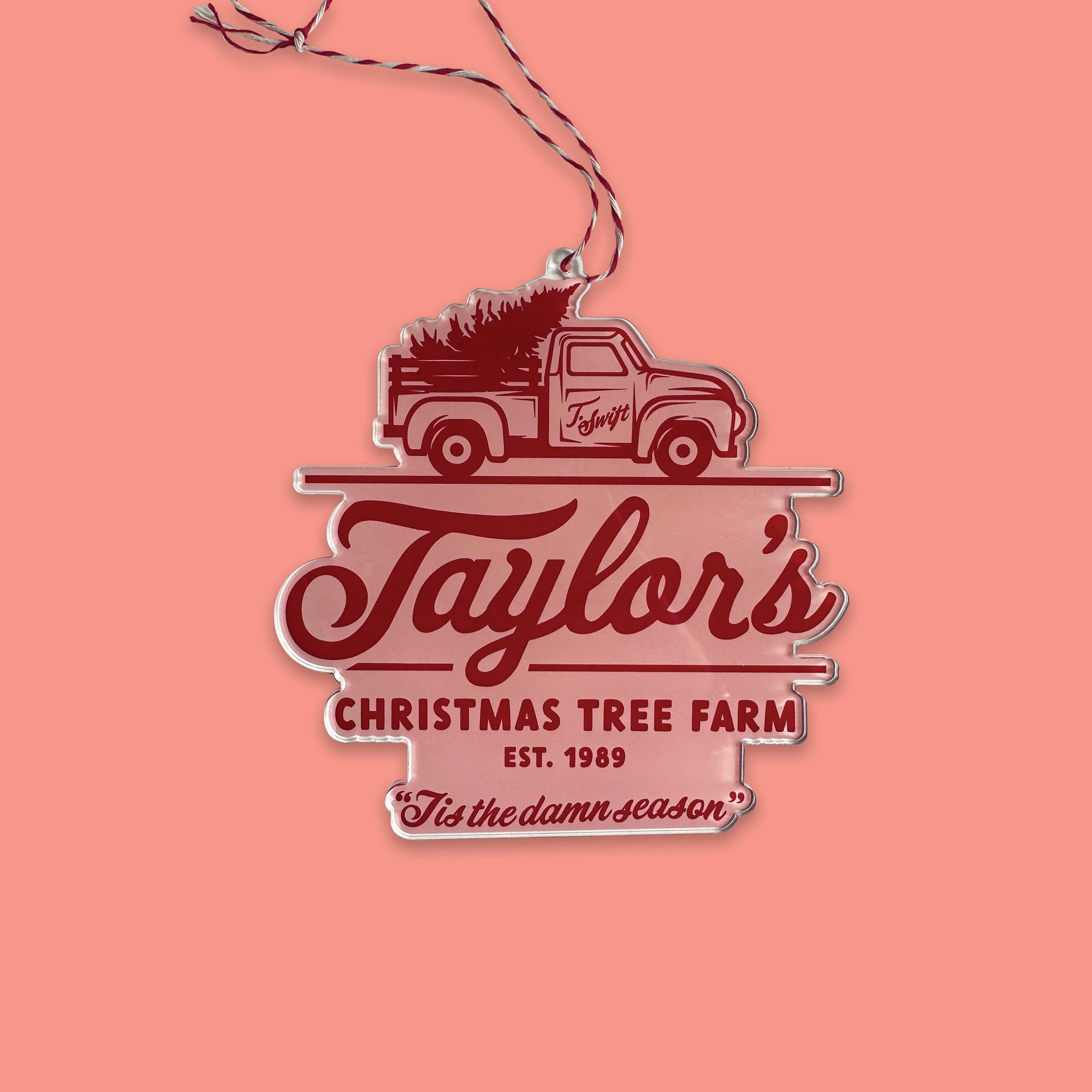 On a coral pink background sits an acrylic ornament with red and white string. This Taylor Swift inspired ornament has a red vintage truck with a tree and it says "Taylor's CHRISTMAS TREE FARM EST. 1989" in hand lettering and it also says "Tis the damn season" in red hand lettering at the bottom. It has a pink background.