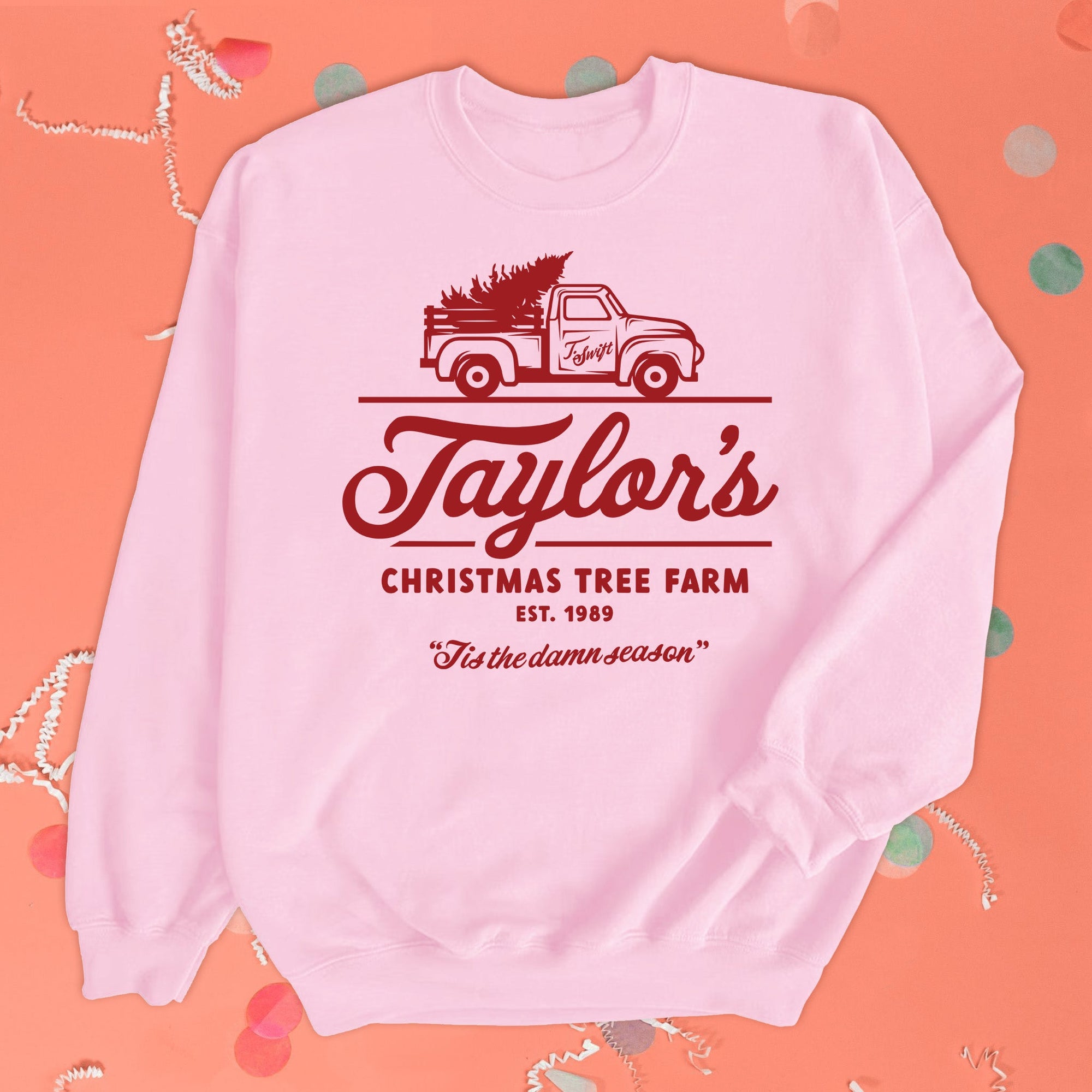 On a coral pink background sits a sweatshirt with white crinkle and big, colorful confetti scattered around. The Taylor Swift inspired pink, crew neck sweatshirt has a red vintage truck with a tree and it says "Taylor's CHRISTMAS TREE FARM EST. 1989" in red hand lettering and it also says "Tis the damn season" in red hand lettering at the bottom.