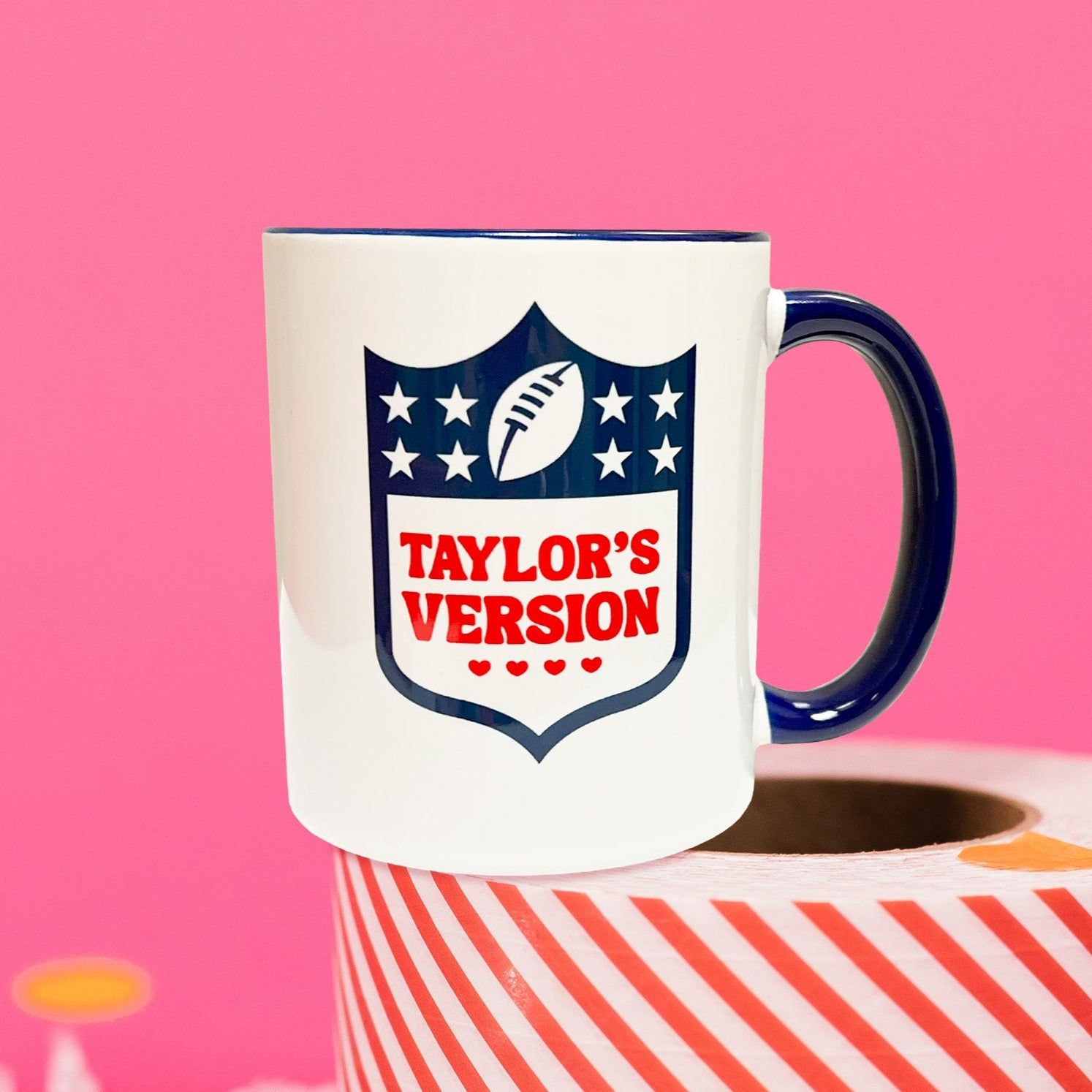 On a hot pink background sits a mug on top of a red and white striped packing tape with white crinkle and big, colorful confetti scattered around. There are mini disco balls. This NFL-Taylor Swift inspired mug has an NFL logo on the front and it says "TAYLOR'S VERSION" with four hearts under it. It is in red.