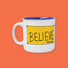 On an orangey-red background sits a mug. This Ted Lasso inspired white mug has a cobalt blue rim and the inside is also cobalt blue. On the front is a sunny mustard yellow rectangle with cobalt blue tape on each corner and it says "BELIEVE" in black, all caps handwritten lettering. It also says "TED LASSO" in all caps bold font.