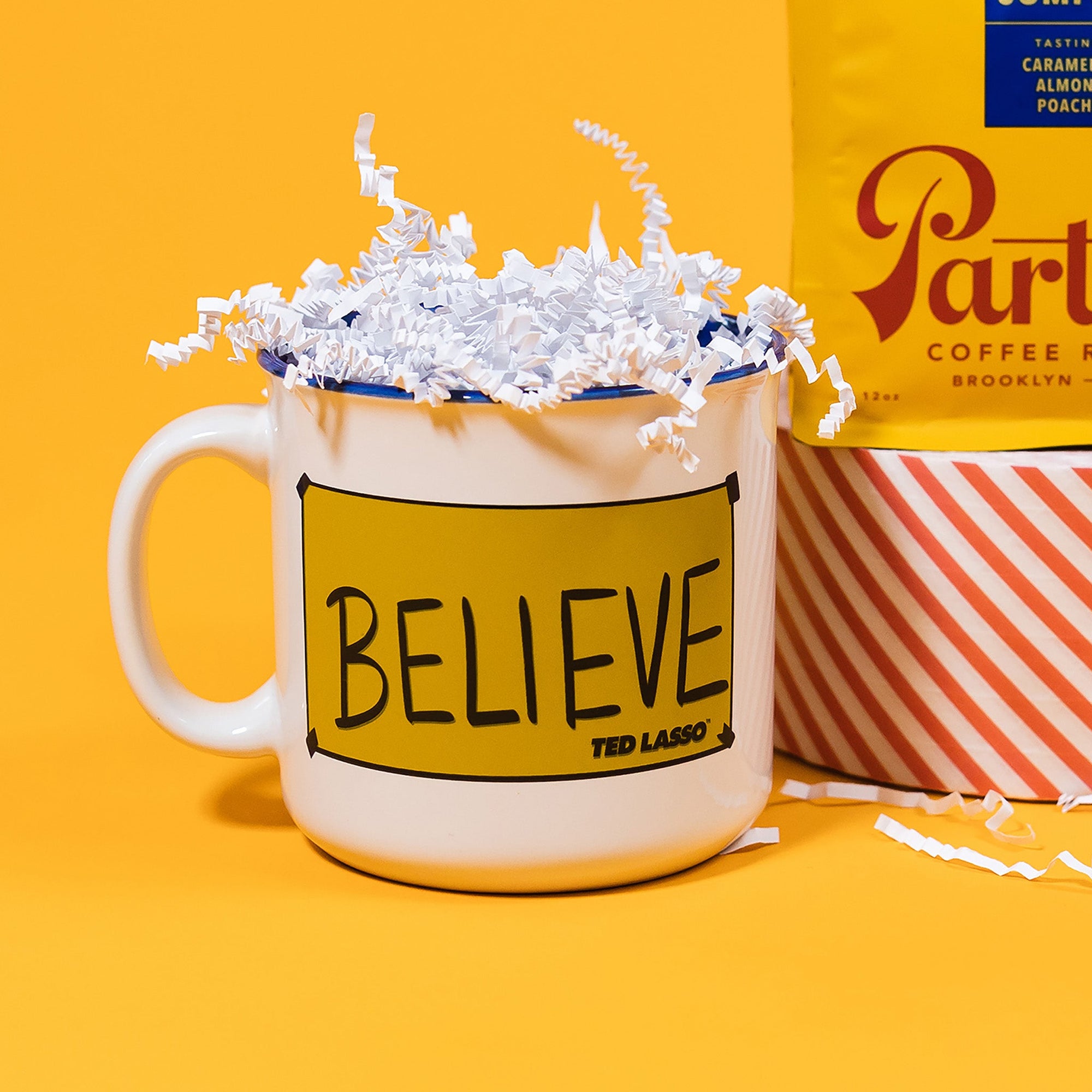 On a sunny mustard background sits a mug with white crinkle in it. This Ted Lasso inspired white mug has a cobalt blue rim and the inside is also cobalt blue. On the front is a sunny mustard yellow rectangle with cobalt blue tape on each corner and it says "BELIEVE" in black, all caps handwritten lettering. It also says "TED LASSO" in all caps bold font.