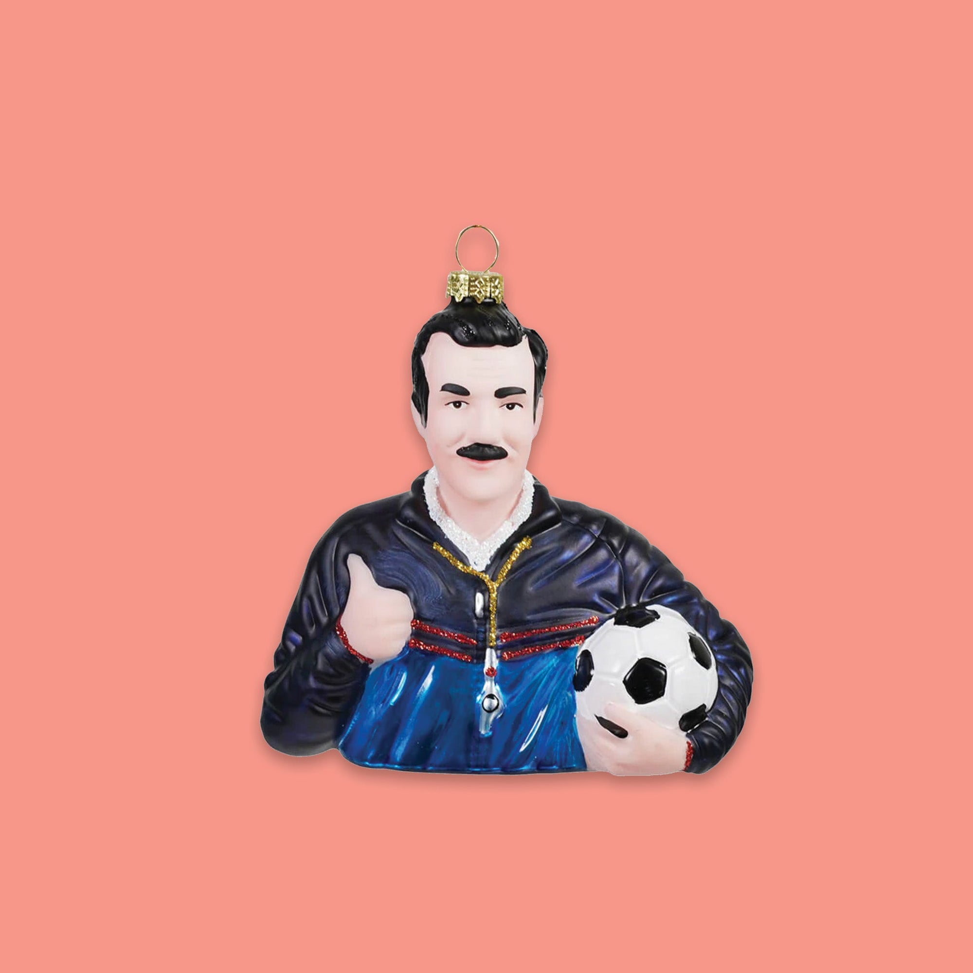 On a coral pink background sits an ornament. This Ted Lasso inspired glass ornament is a bust of Ted Lasso himself wearing a coach's jacket and whistle, doing a thumb's up and carrying a soccer ball.