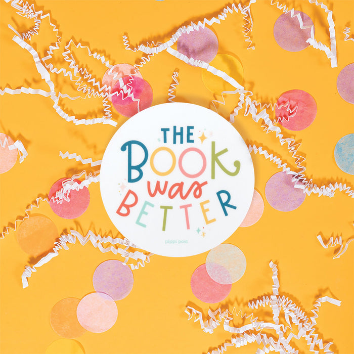 On a sunny mustard background is a sticker with white crinkle and big, colorful confetti scattered around. This Harry Potter inspired round white sticker has colorful lettering on it. It says "THE BOOK WAS BETTER." 3"x3"