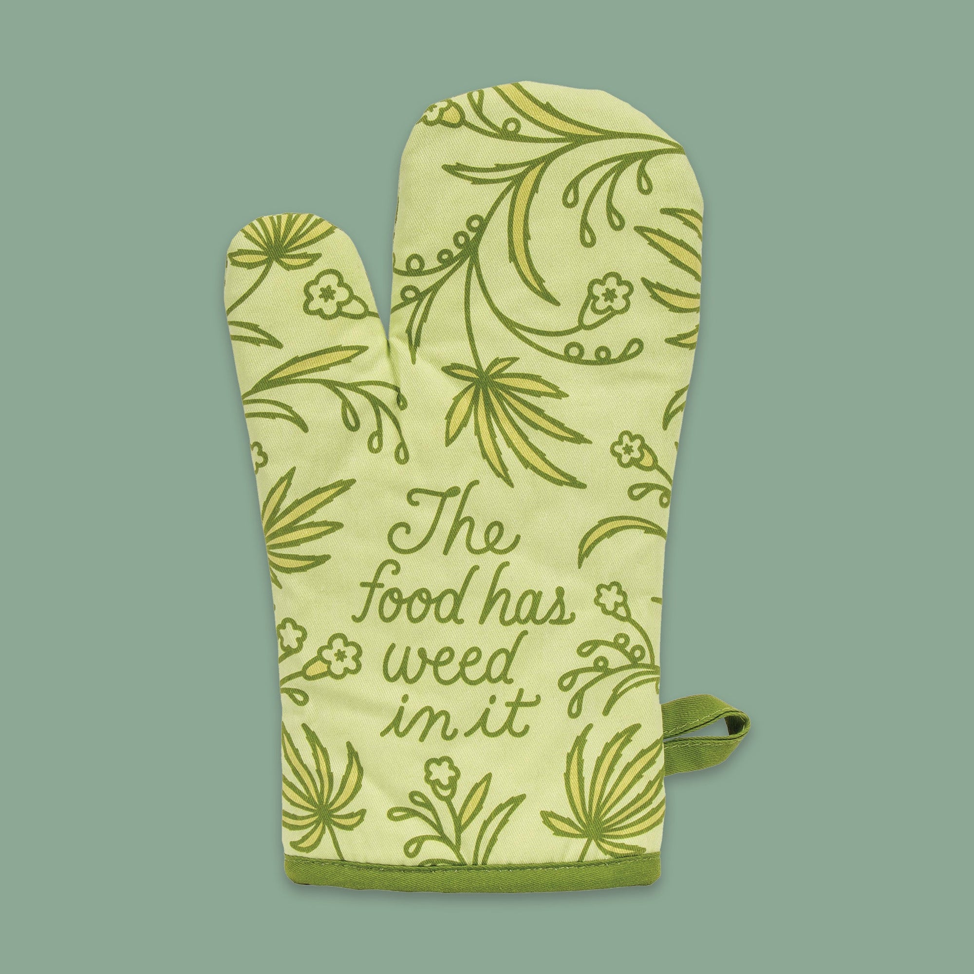On a moss green background sits an oven mitt. The front of this celery green oven mitt has illustrations of green marijuana leaves and flowers on it and it says "The Food has weed in it" in green handwritten script lettering.