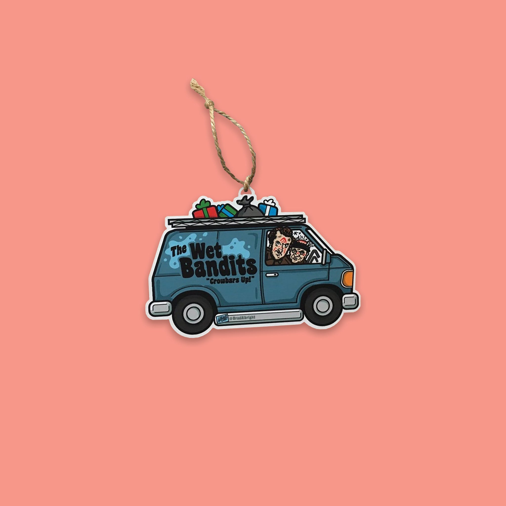 On a coral pink background sits an ornament. This Home Alone themed wood ornament is of The Wet Bandits blue van with Harry and Marv sitting in it. There are presents on top of the van and there is a twine rope tied to the ornament. 