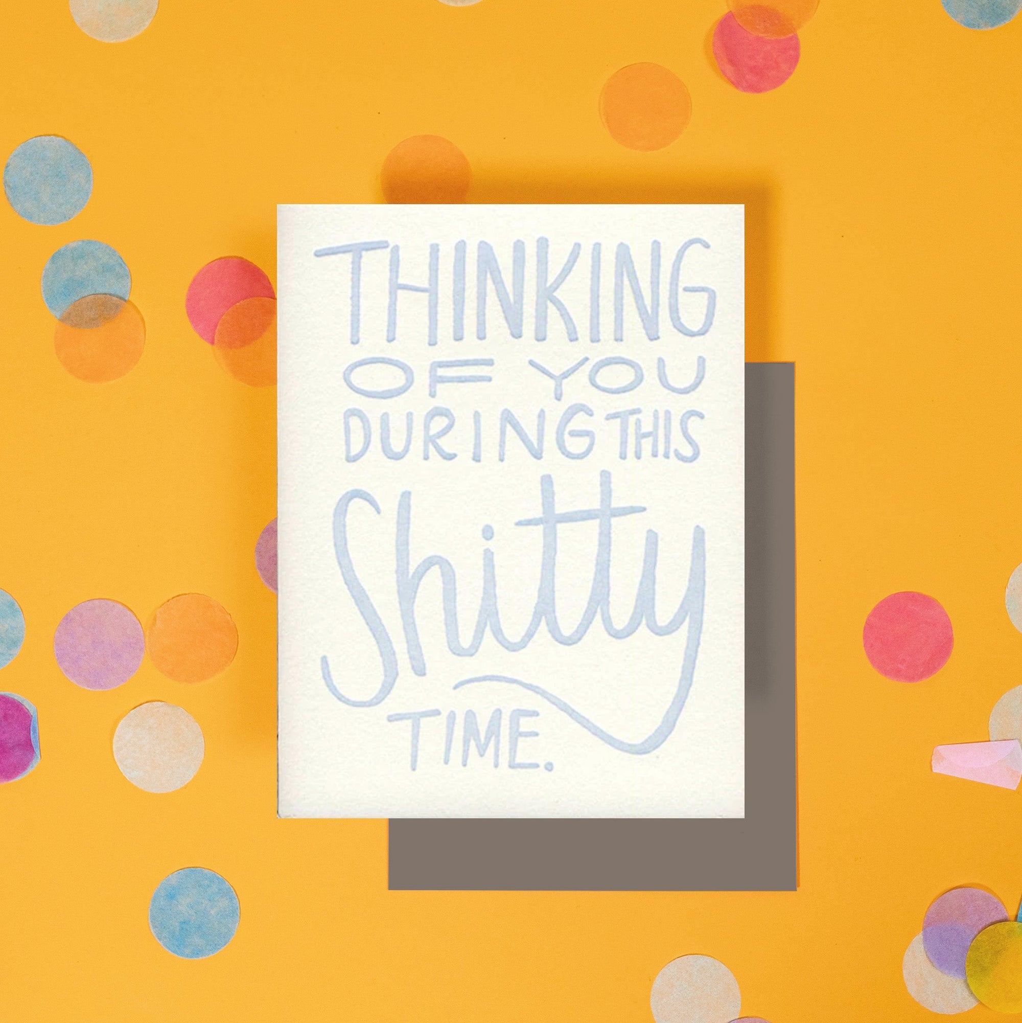 On a sunny mustard background is a greeting card and envelope with big, colorful confetti scattered around. The white greeting card says "Thinking of you during the shitty time." in a light blue, handwritten letterpress lettering. A taupe brown envelope sits under the card.