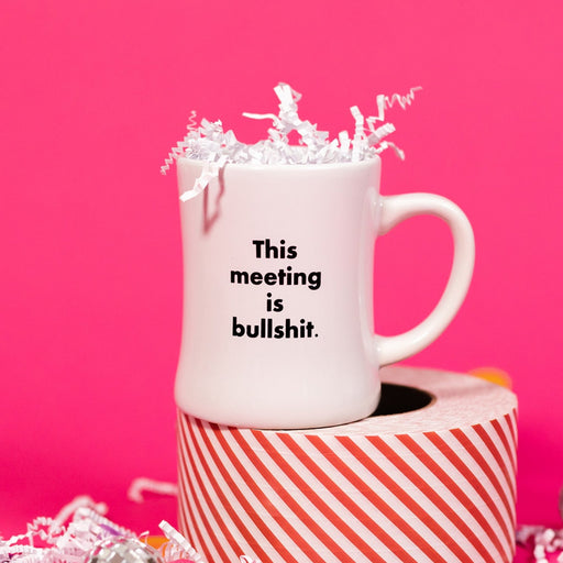 On a hot pink background sits a mug with white crinkle and big, colorful confetti scattered around. This white, diner style mug says "This meeting is bullshit." in black, block font. It has white crinkle in it and sits atop a red and white striped roll of tape. 