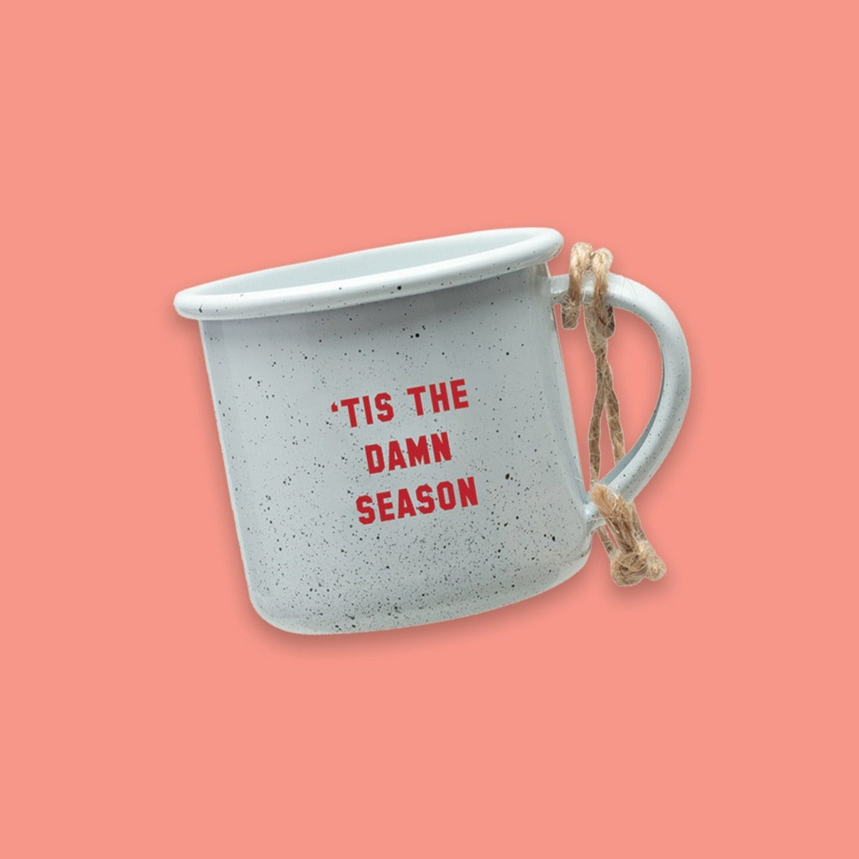 On a coral pink background sits a white mini campfire mug with black specks. It says "'TIS THE DAMN SEASON" in red collegiate lettering. It has twine on the handle.
