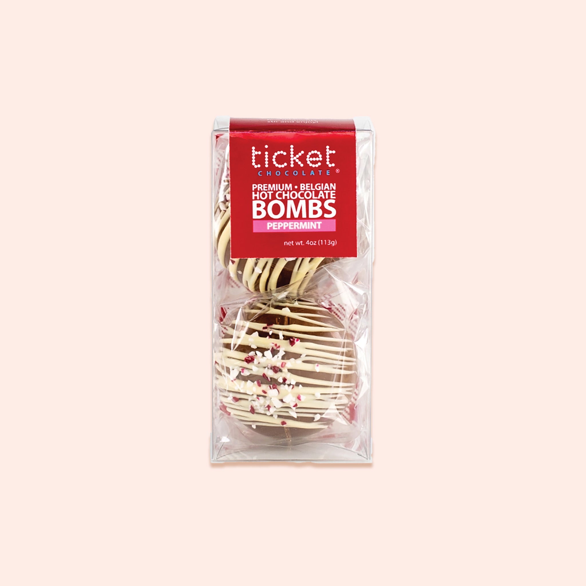 On a light pink background sits a clear package with a red label. Inside the package are two chocolate covered hot chococlate bombs with white chocolate drizzled on top and crumbled peppermint. On the label it says "ticket CHOCOLATE PREMIUM • BELGIAN HOT CHOCOLATE BOMBS PEPPERMINT." net wt. 4oz (113g)