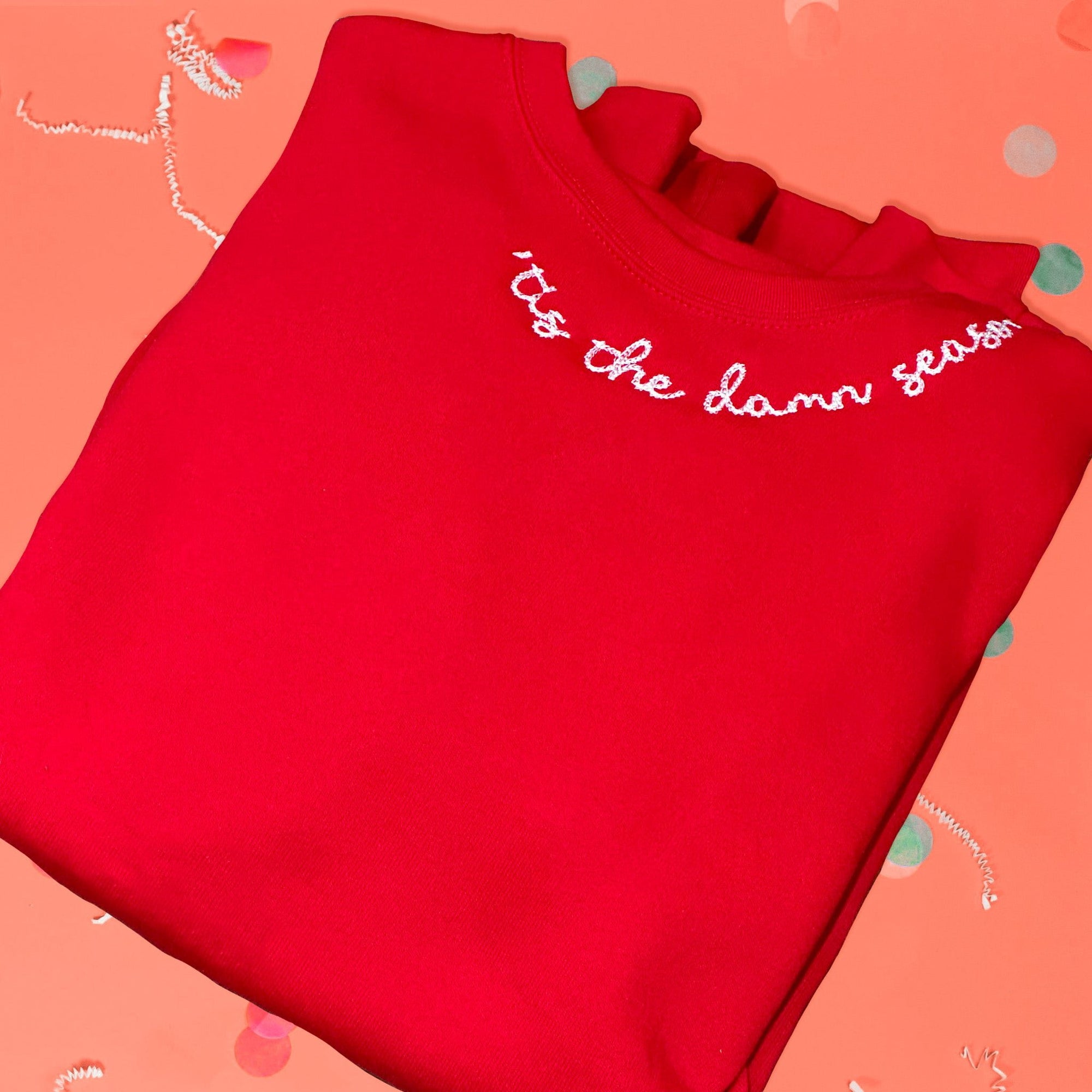 On a pink coral background sits the front of a sweatshirt with white crinkle and big, colorful confetti scattered around. This Taylor Swift Inspired crewneck red sweatshirt has white chain stitched script lettering around the neck and it says "'tis the damn season."