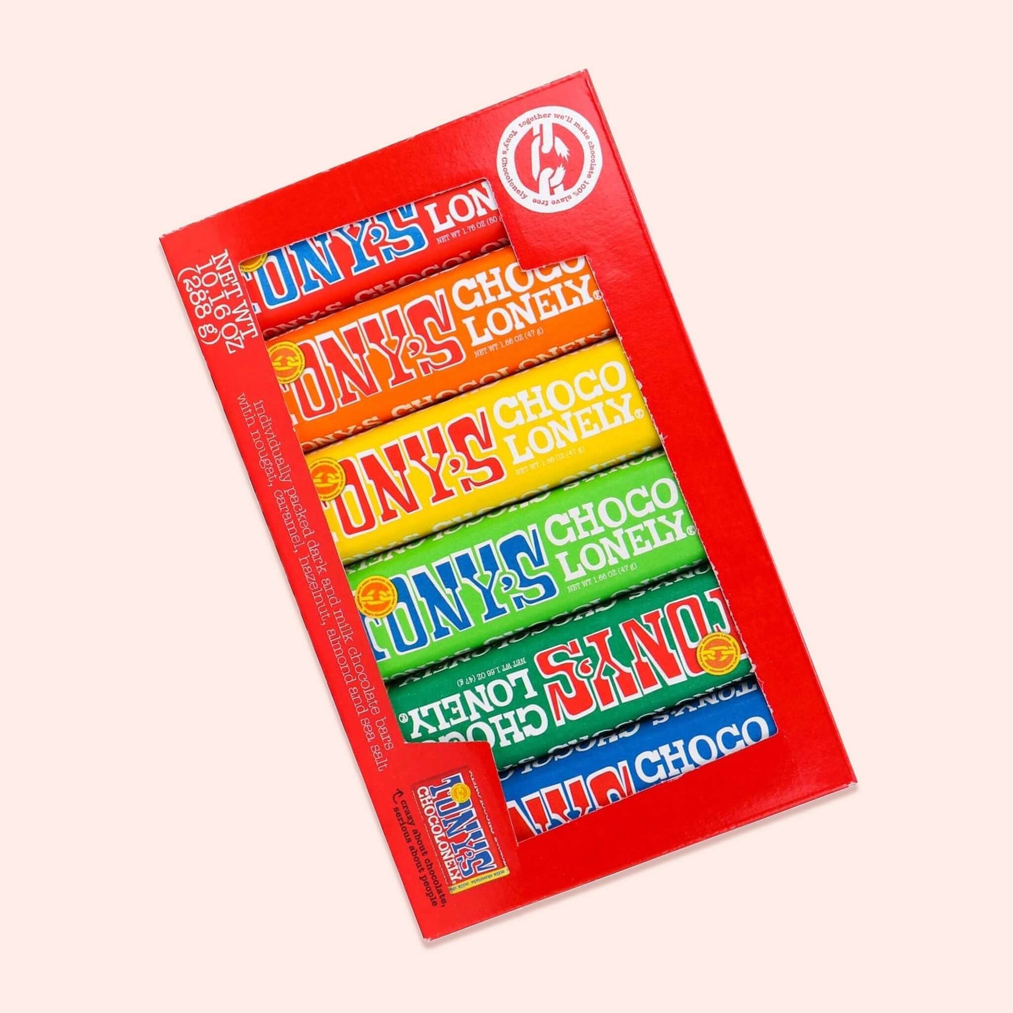 On a light pink background sits a red box with a see-through window on the front. It is filled with colorful packaged chocolate bars. There are red, orange, yellow, lime green, dark green, and blue wrapped bars and they all say "TONY'S CHOCOLONELY." It also says "individually packed dark and milk chocolate bars with nougat, caramel, hazelnut, almond and sea salt." NET WT 10.16 OZ (288g)