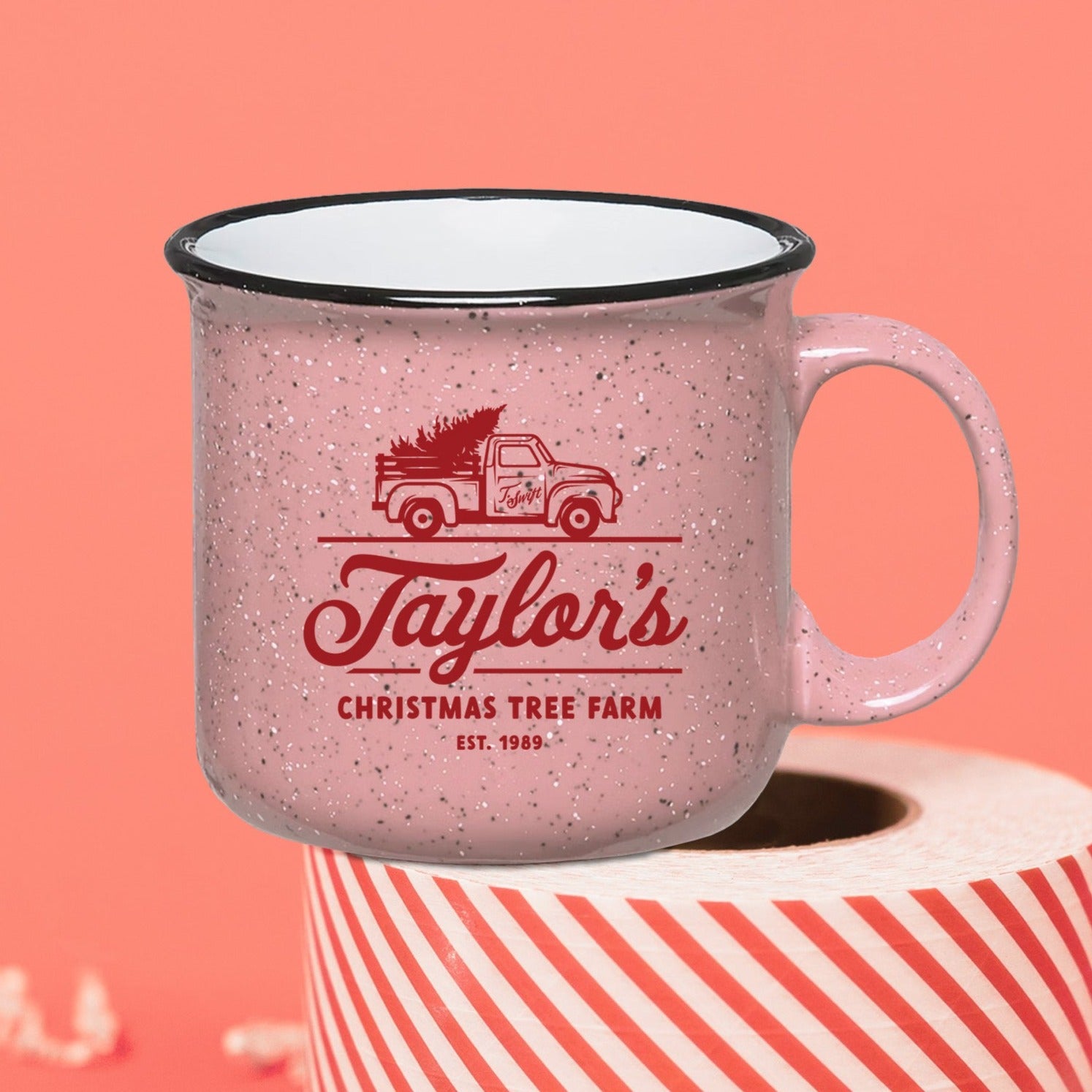 On a coral pink background sits a coffee mug on top of a red and white striped packing tape with white crinkle and big, colorful confetti scattered around. There are mini disco balls.  This Taylor Swift inspired pink campfire mug has a red vintage truck with a tree and it says "Taylor's CHRISTMAS TREE FARM EST. 1989" in red hand lettering and it also says "Tis the damn season" in red hand lettering at the bottom.